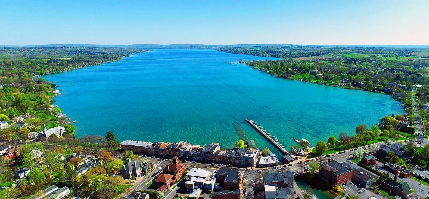 Aerial view of a large blue lake surrounded by a town with green trees and buildings, with a road and dock on the shoreline.
