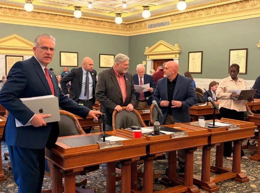 Group of professionals in suits and business casual attire in a formal meeting room with wooden desks, microphones, and framed documents on the walls.