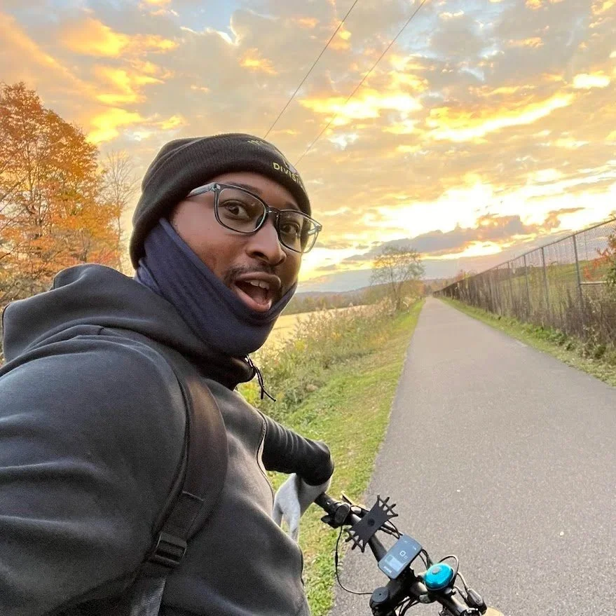 A man taking a selfie during sunset while riding a bicycle on a paved trail surrounded by trees with autumn foliage.