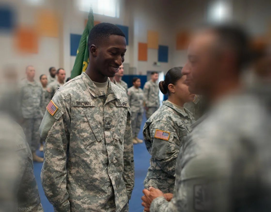 A smiling African American U.S. Army soldier in military uniform shaking hands with a foreign officer in military uniform at a military ceremony, with other soldiers standing in formation in the background.