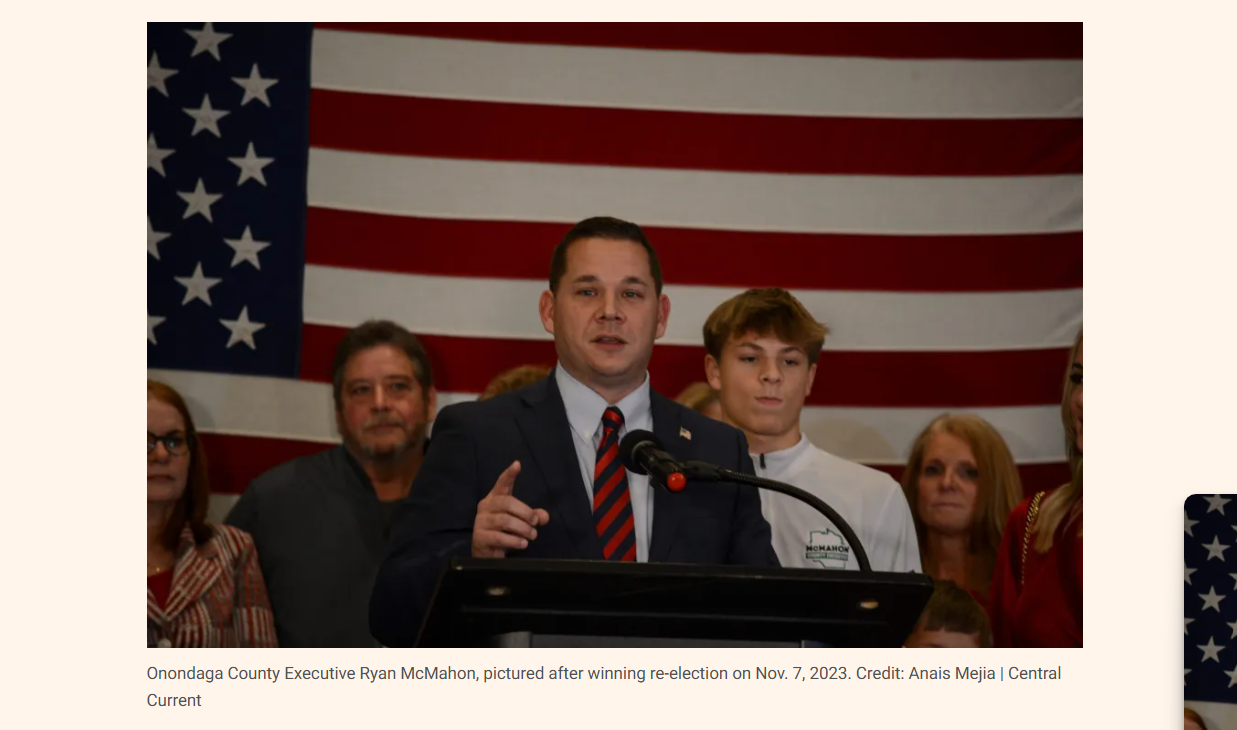Onondaga County Executive Ryan McMahon speaking at a podium after winning re-election, with a large American flag in the background, and several people standing behind him.