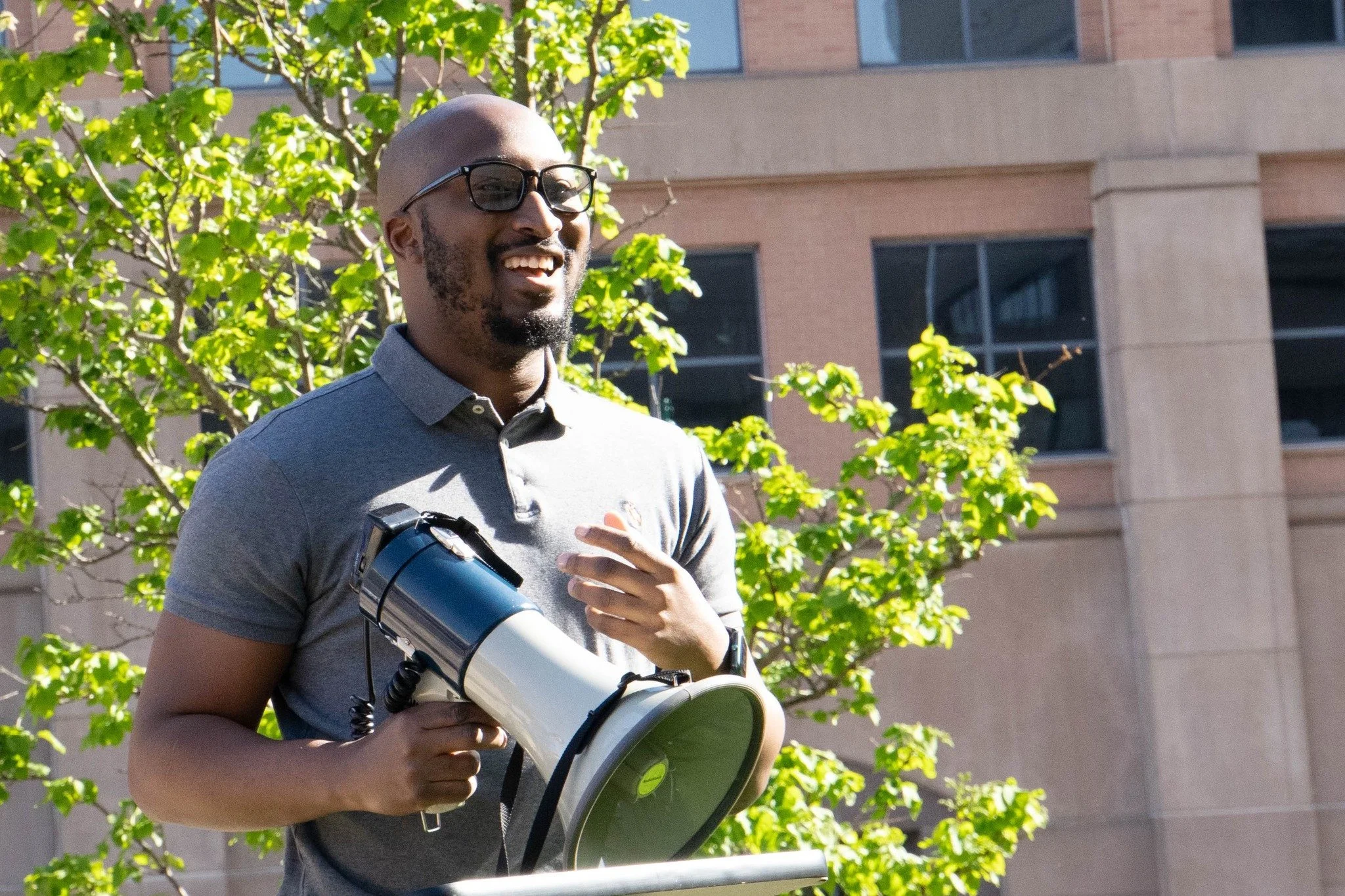 A man with glasses, a beard, and a shaved head holding a megaphone and a notebook outdoors near green trees and a building.