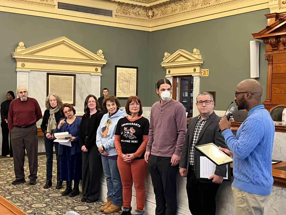 A group of eight diverse people are standing in a line in a formal room, possibly a city hall or courtroom, listening to a man in a blue sweater who is speaking into a microphone and holding a document. The room has ornate ceiling molding and framed documents on the wall.