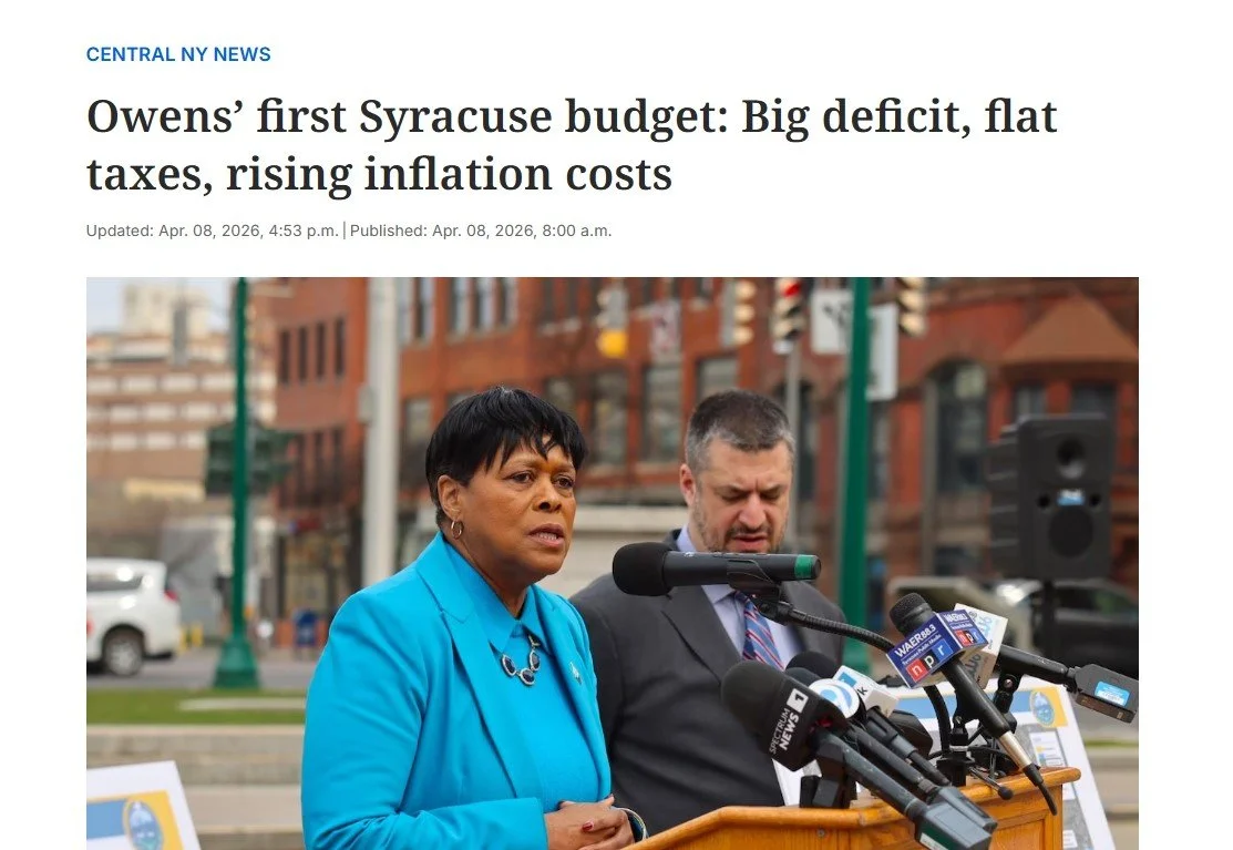 Two officials, a woman in a blue blazer and a man in a suit, speaking at a press conference outdoors with microphones and banners about a Syracuse budget issue.