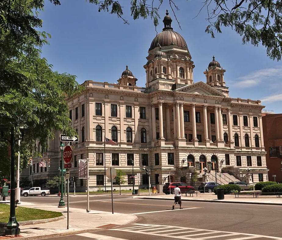 A historic government building with a large dome and columns, situated at a street corner with traffic signs and trees.