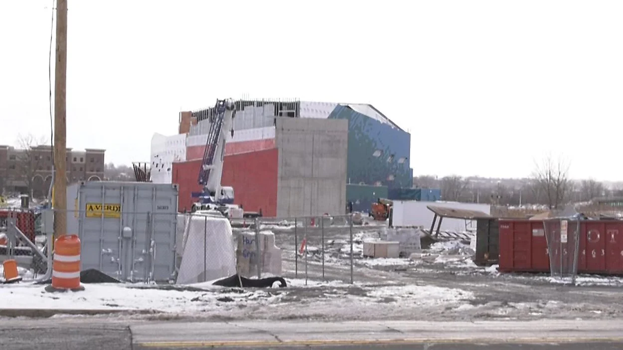 Construction site in winter with a partially built modern building, a crane, construction materials, safety barriers, and snow on the ground.