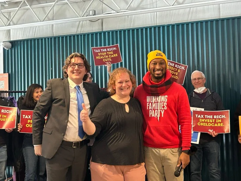 Group of people at a rally or protest holding signs that say "Tax the Rich" and "Stop the Health Care Cuts." The group appears happy and supportive of their cause.