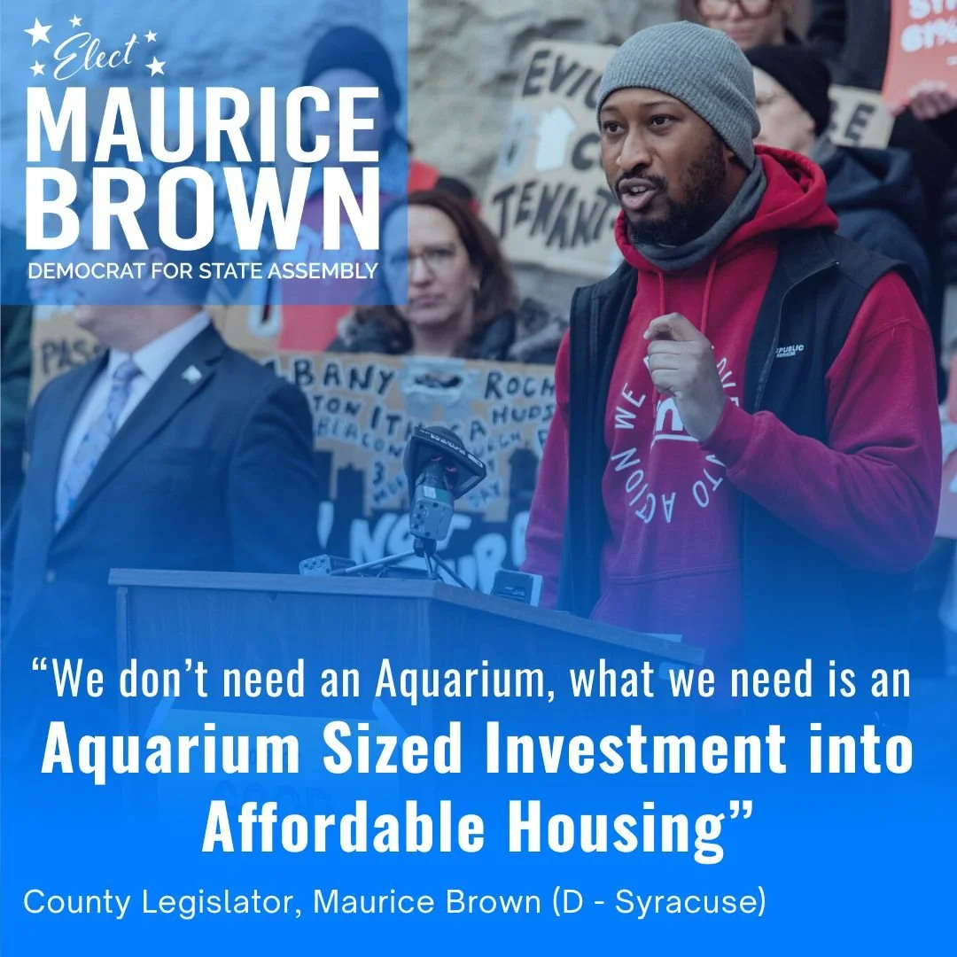 Maurice Brown, a Black man in a gray beanie and red hoodie, speaking at a rally with a podium, surrounded by protesters holding signs, advocating for investment in affordable housing.