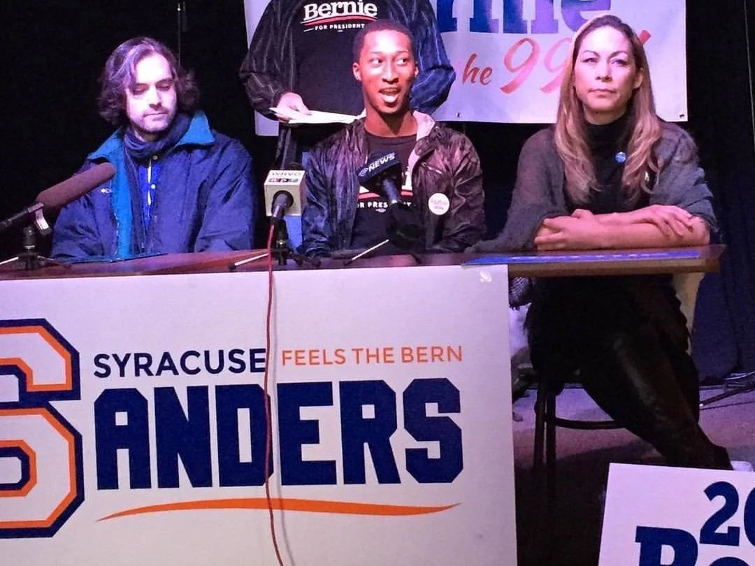 Three people sit at a table with microphones, at a political event supporting Bernie Sanders. The middle person is speaking, while the person on the left appears to be listening and the person on the right has arms crossed. A banner and signs are visible in the background.