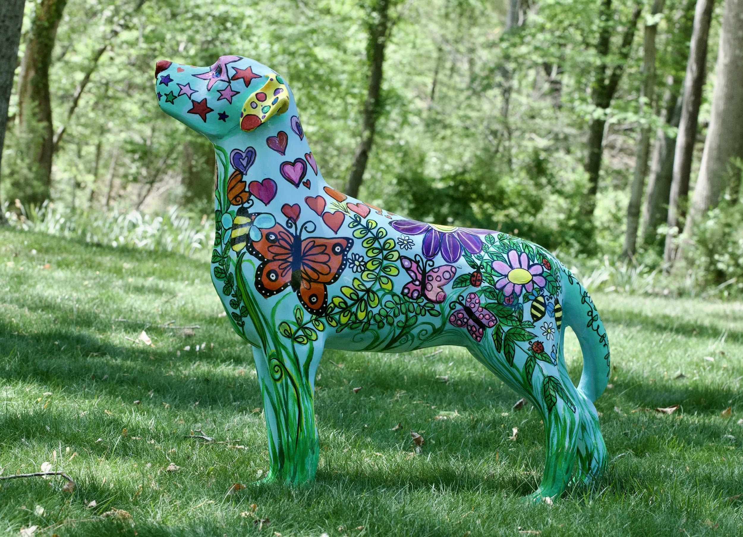 Colorful painted sculpture of a dog outdoors in a wooded area with green trees and grass.