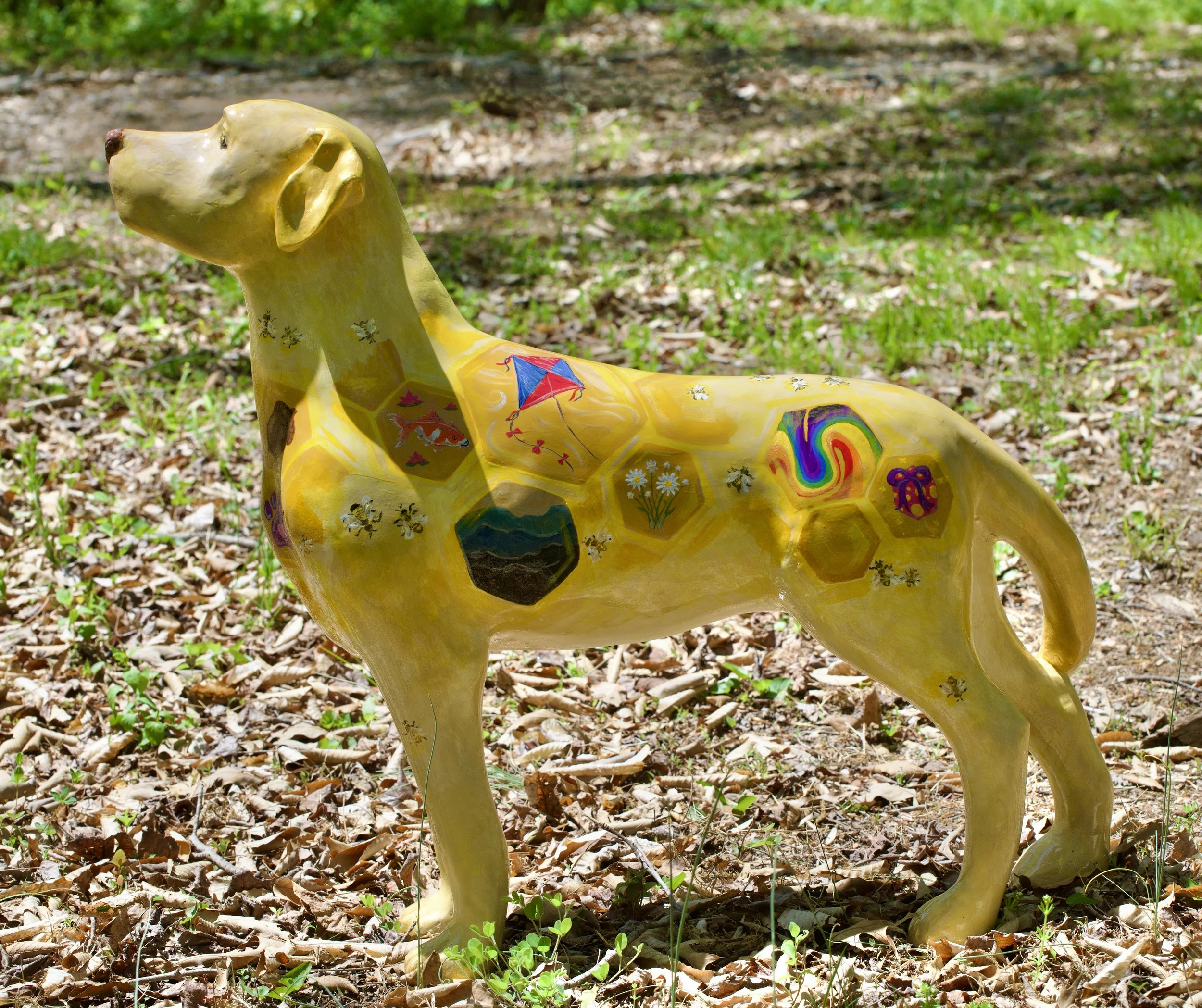 Colorful ceramic sculpture of a dog with painted designs, sitting outdoors on a ground covered with dry leaves and green plants.