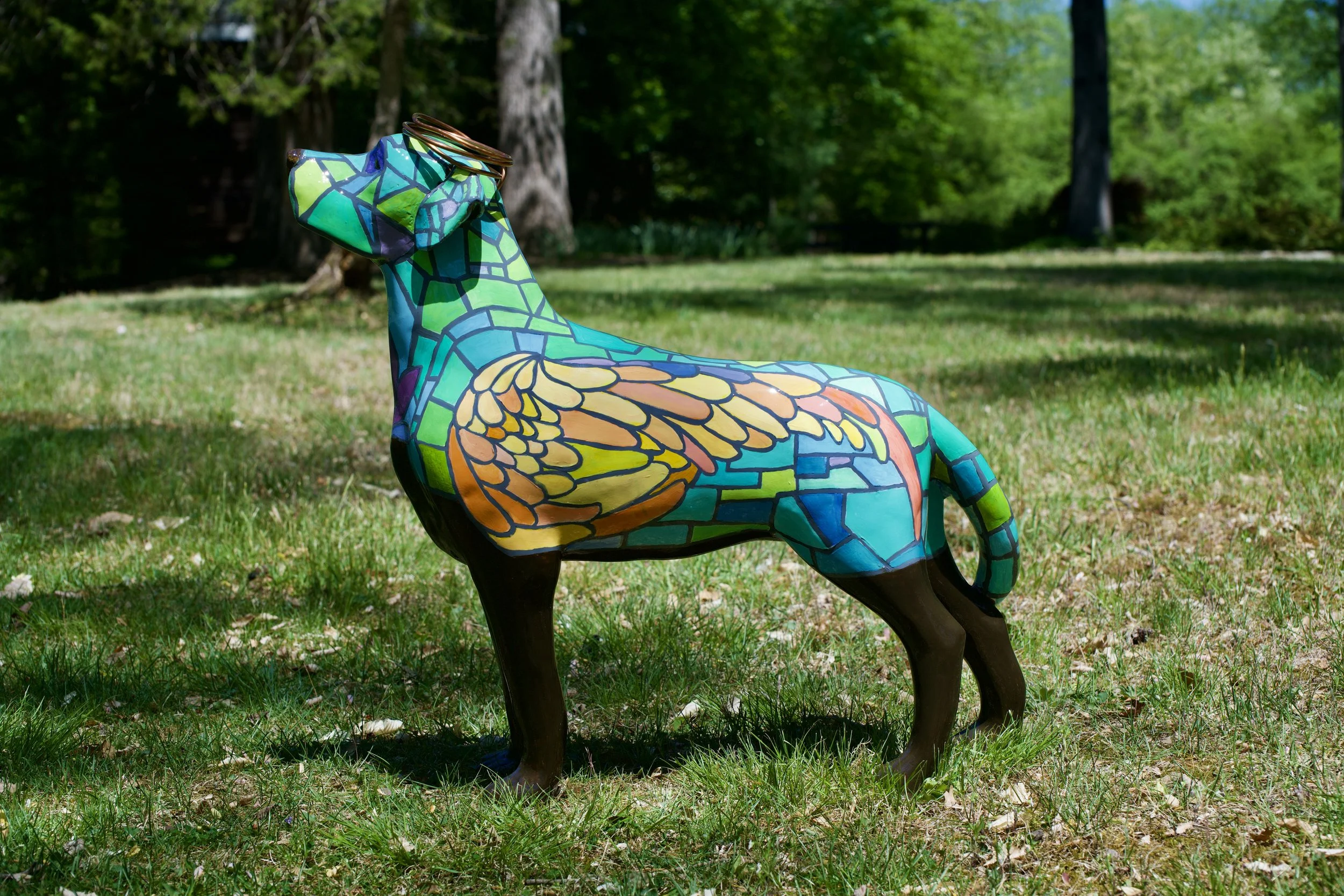 Decorative horse sculpture painted with a colorful mosaic pattern in green, blue, yellow, orange, and purple, situated outdoors on grass with trees in the background.