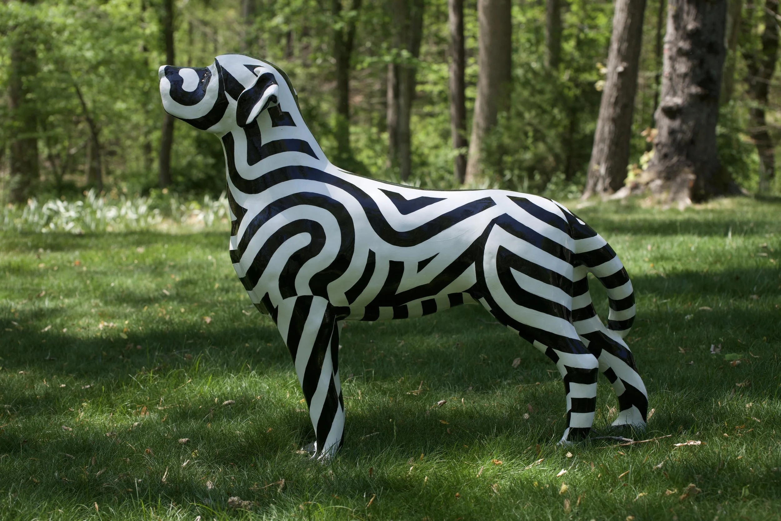 A life-sized sculpture of a zebra painted with black and white zebra stripe patterns, situated in a grassy area surrounded by trees.