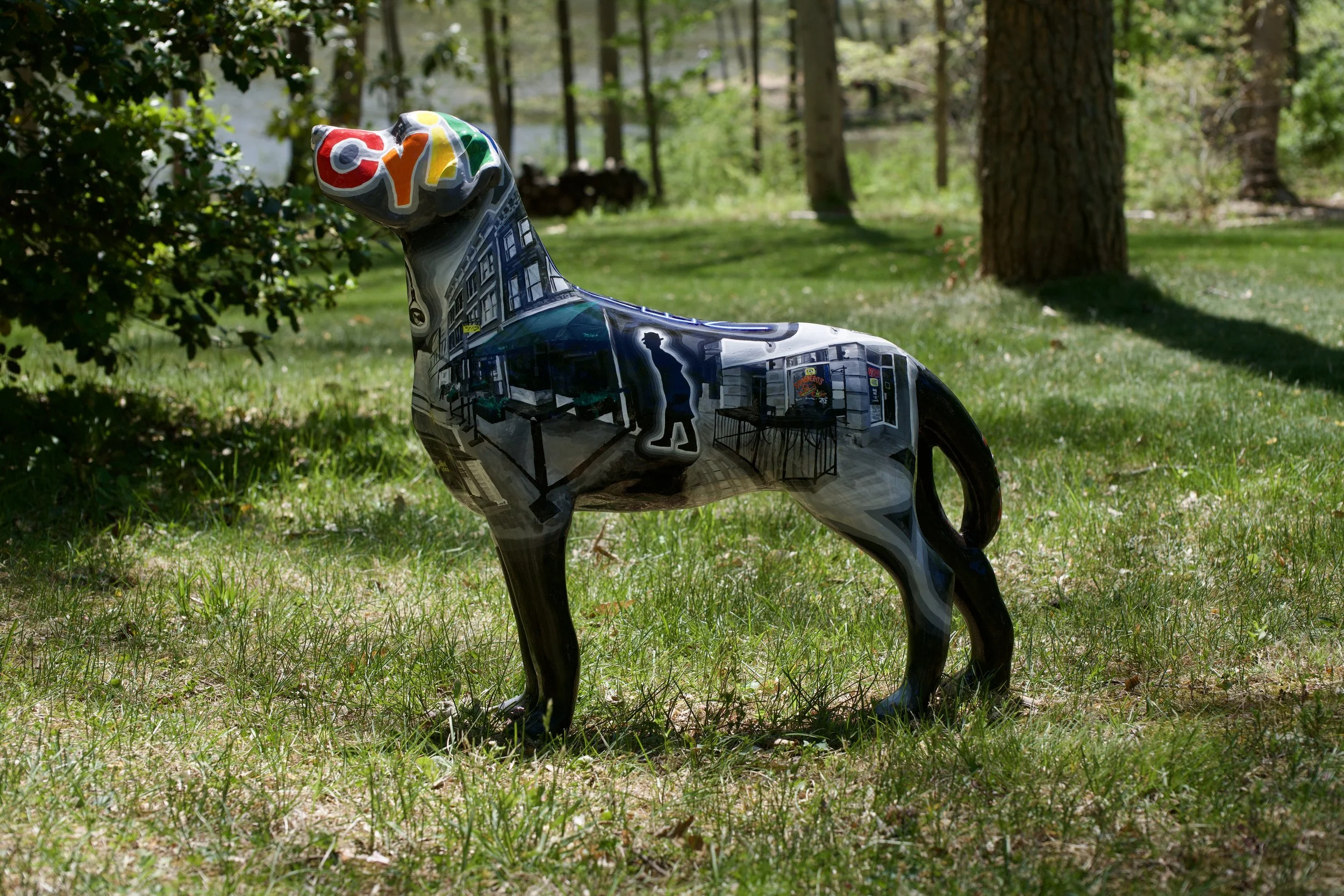 Balloons shaped like a dog with colorful decorations are placed outdoors on grass in a park, with trees and sunlight in the background.