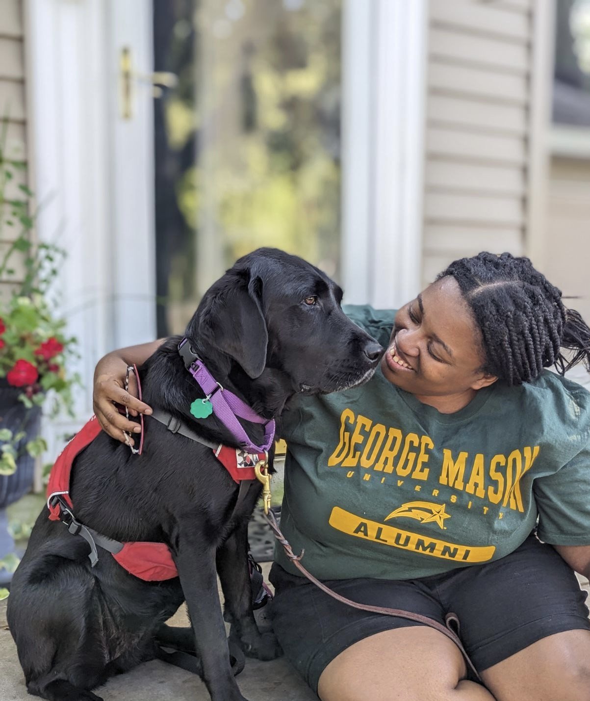 Person sitting with a black dog wearing a red harness, smiling at each other. The person is wearing a green George Mason University Alumni t-shirt. There's a potted plant with red flowers in the background.