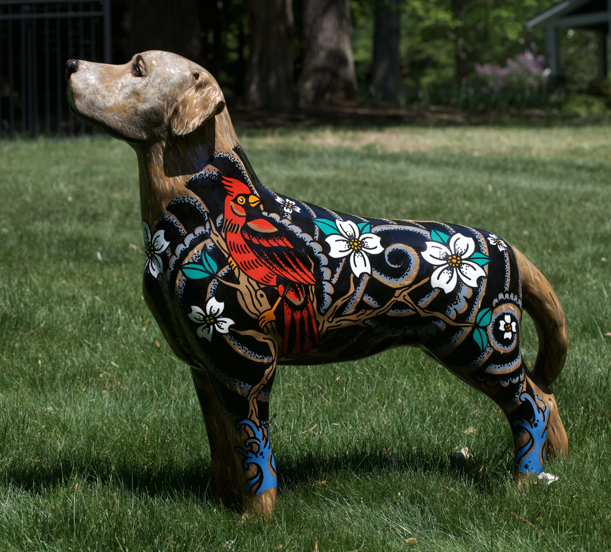 A sculpture of a dog painted with colorful, intricate patterns including flowers, a cardinal bird, and abstract designs, situated outdoors on a grassy area with trees in the background.