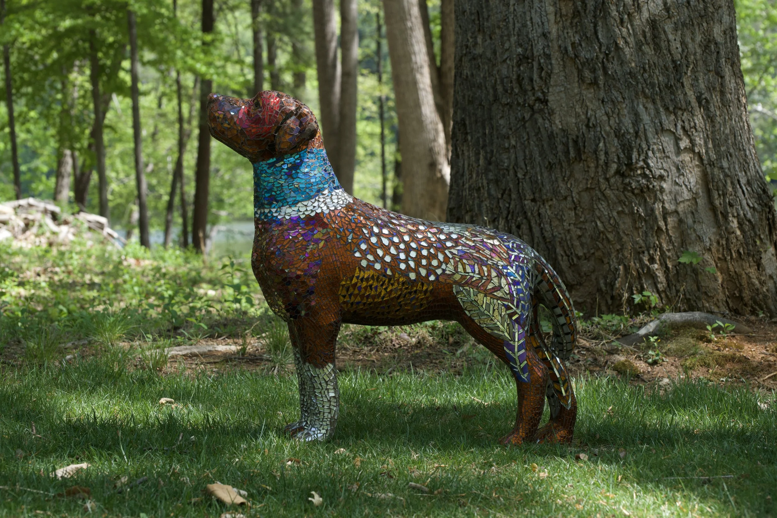 A mosaic sculpture of a dog standing on grass in a forested area with trees in the background.