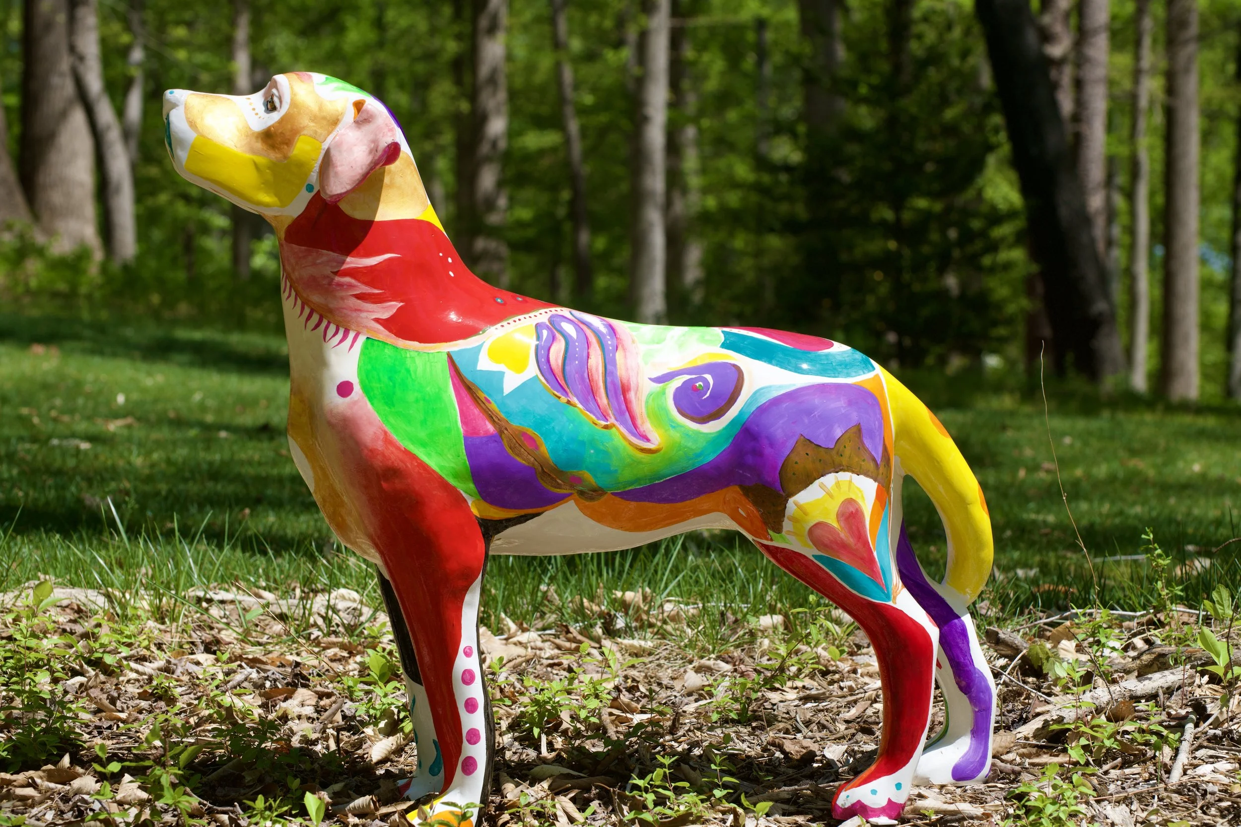 Colorful painted dog sculpture in a forest setting with trees and grass.