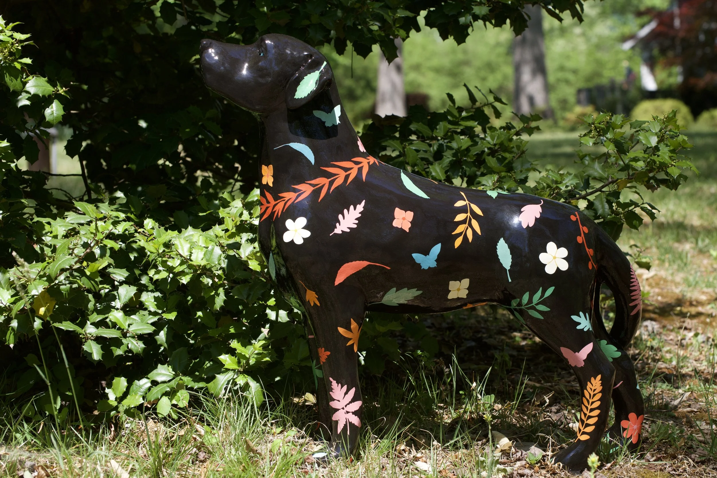 Painted black sculpture of a dog with colorful floral and butterfly patterns, situated among green bushes and trees outdoors.