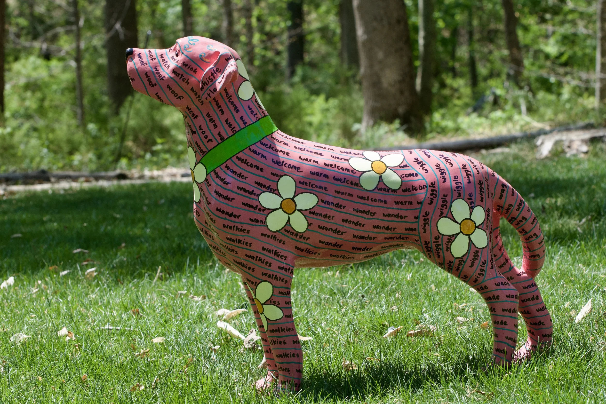 Colorful inflatable giraffe with a floral and text pattern, standing on grass in a forested area.