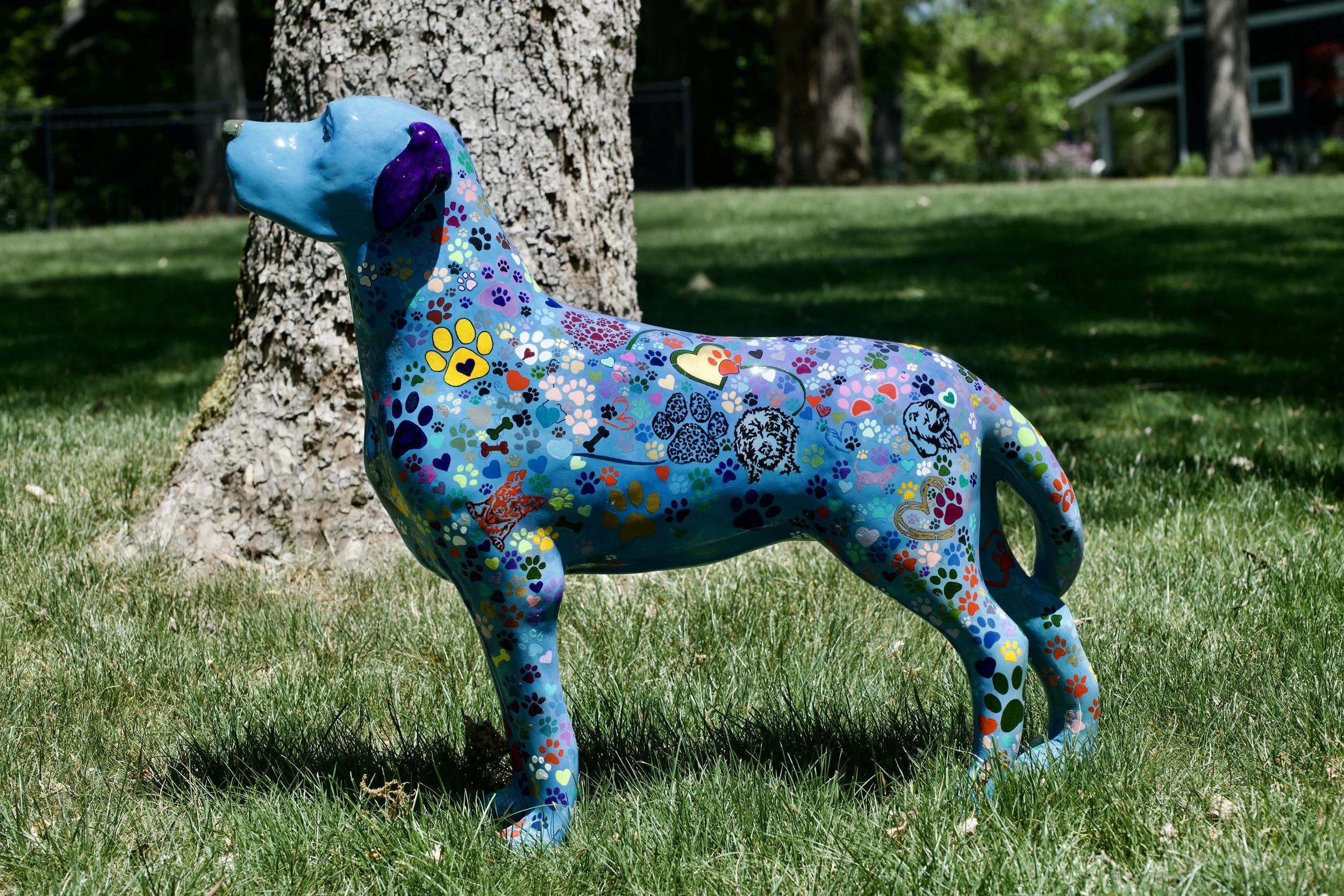 Colorful dog sculpture covered in multicolored paw print patterns, standing on grass next to a tree in a park.