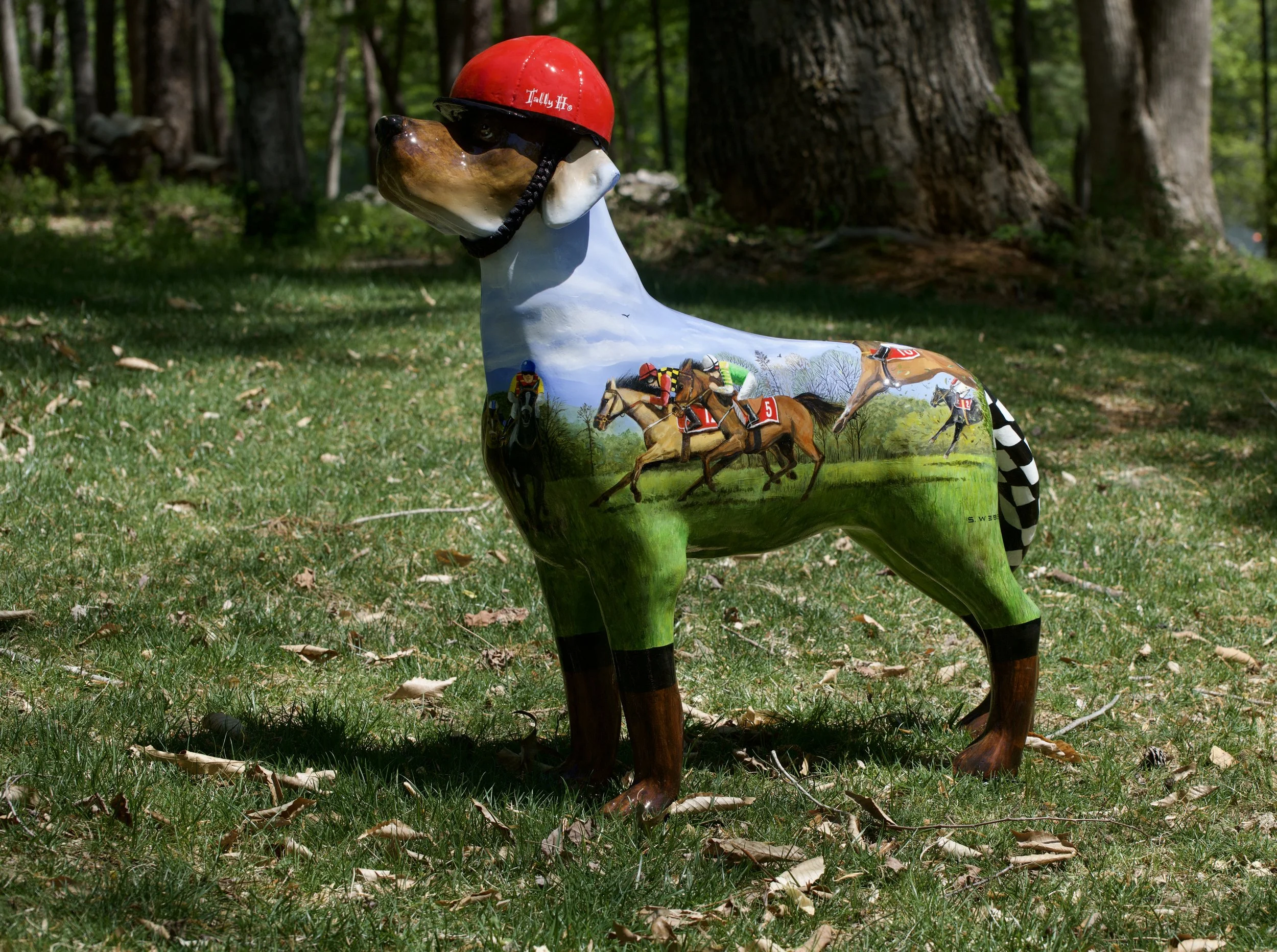 Decorative horse sculpture with a jockey helmet, painted with images of horse racing, placed on a grassy area in a wooded park.