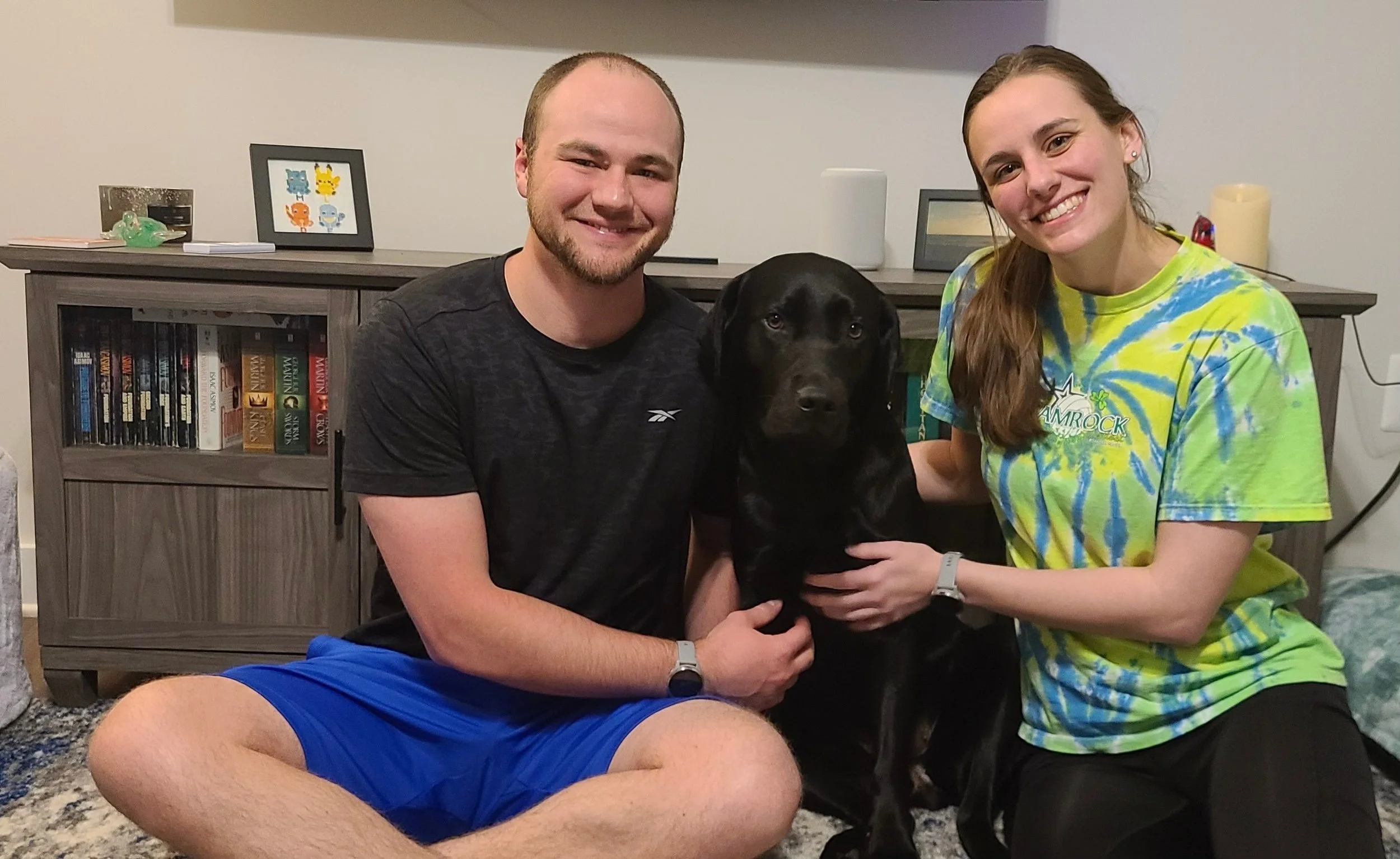 Two young adults, a man and a woman, are smiling as they pose with a black dog indoors. The man is wearing a dark shirt and blue shorts, while the woman is wearing a tie-dye T-shirt. They are sitting on the floor in front of a bookshelf. The woman is holding the dog, and both appear happy.