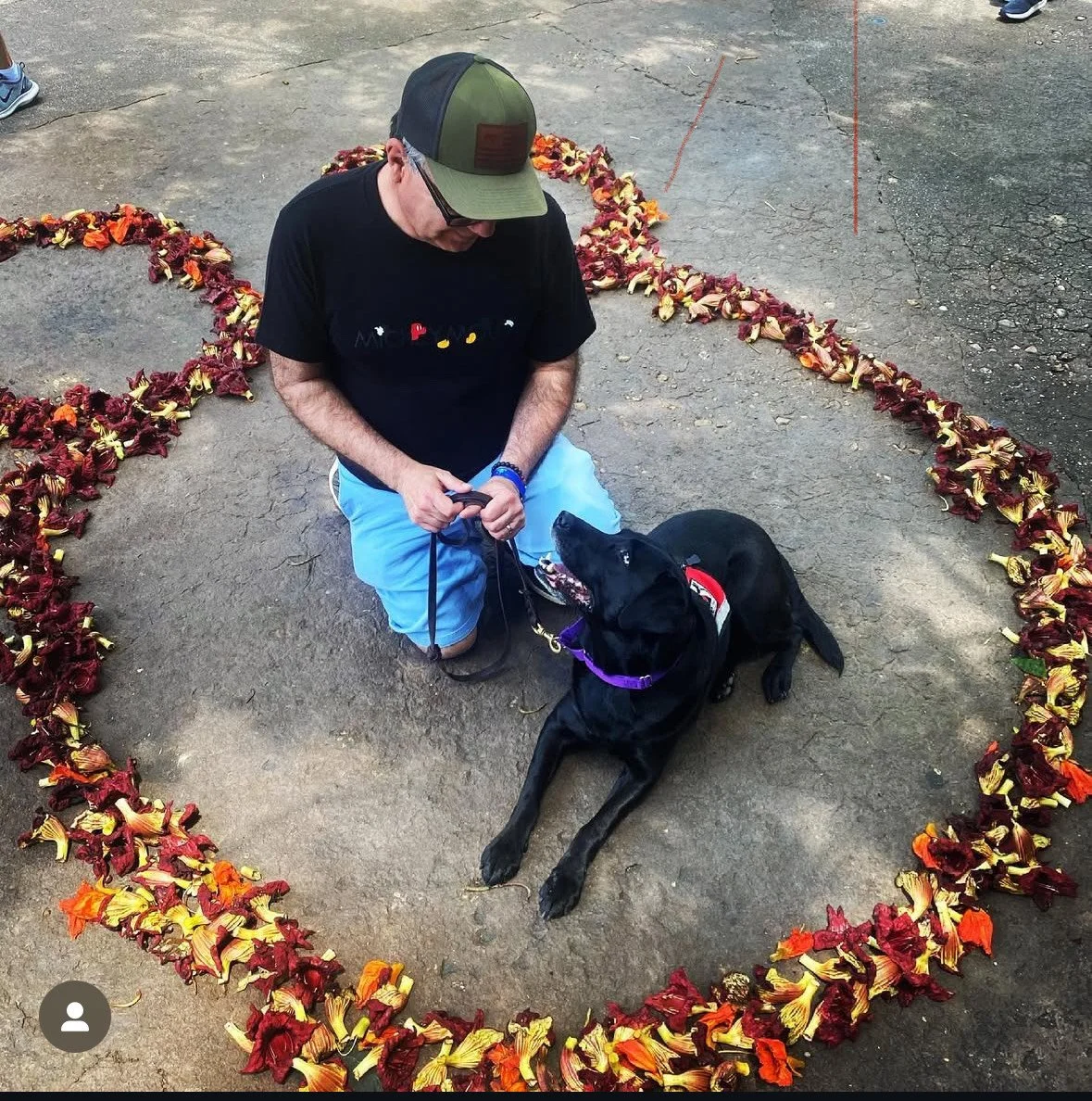 A man kneeling on the ground with a black dog, both inside a heart-shaped outline made of red, orange, and yellow leaves. The man wears a baseball cap, sunglasses, and a black t-shirt, and the dog is lying and looking up at the man.