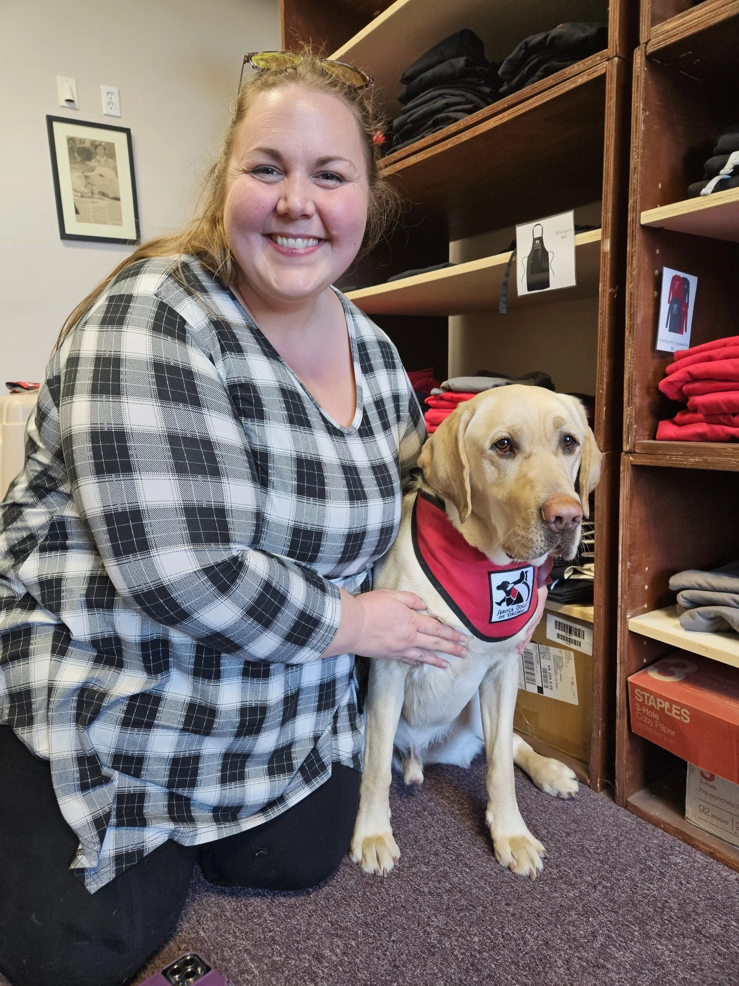 A smiling woman kneeling beside a service dog in an indoor setting, with shelves of clothing in the background.