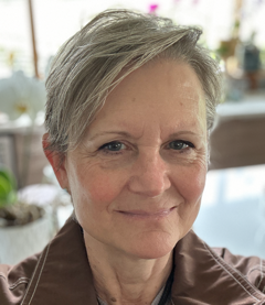 Close-up of a woman with short gray hair smiling indoors, with a blurred background of a room with large windows and plants.
