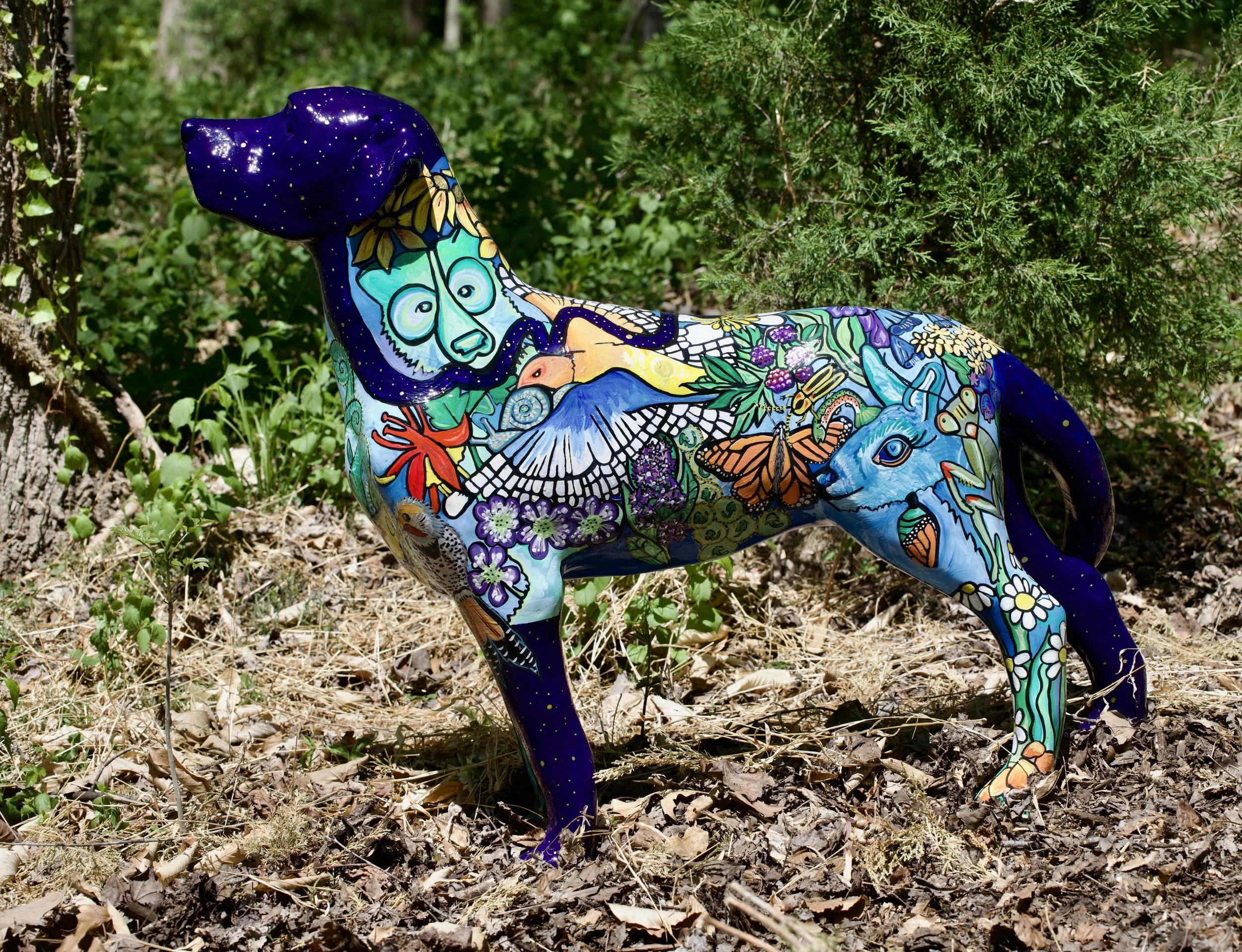 Decorated ceramic dog sculpture with colorful paintings of flowers, butterflies, and various nature motifs, positioned outdoors among trees and ground cover.