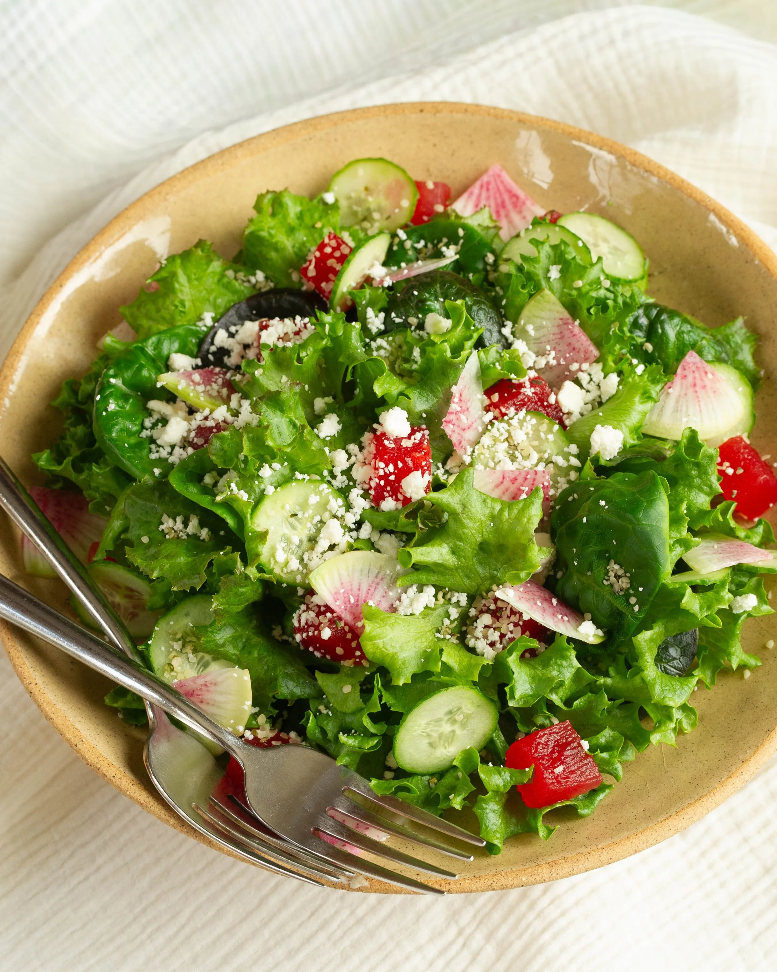 A fresh vegetable salad with lettuce, cucumbers, radishes, red bell peppers, and crumbled cheese in a beige bowl with a metal fork, on a light-colored wooden surface.