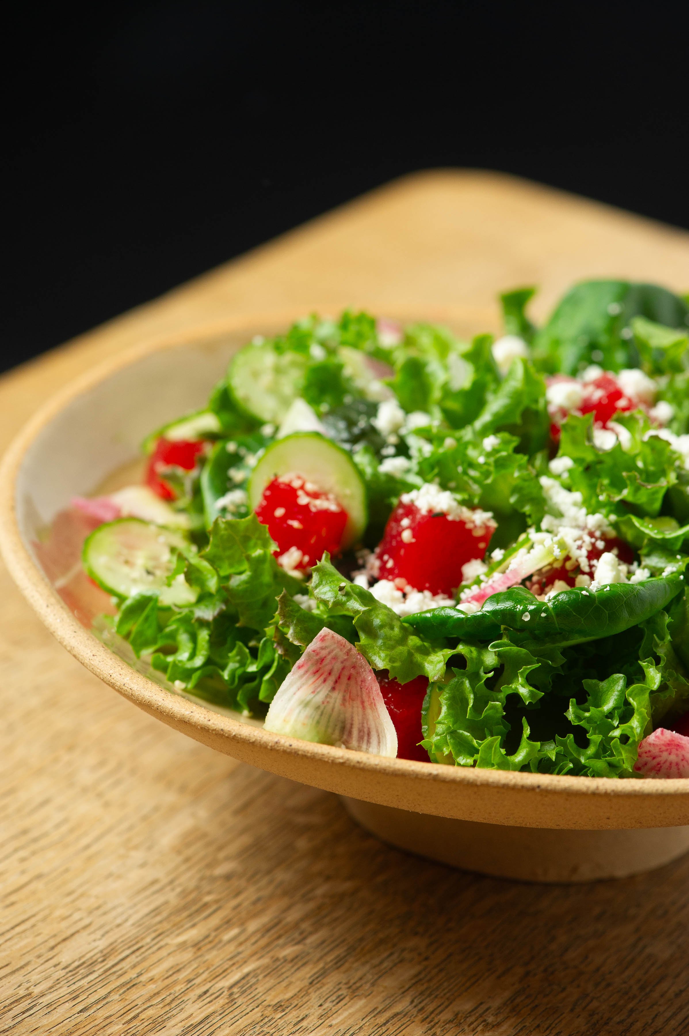 A bowl of fresh salad with leafy greens, sliced cucumbers, cherry tomatoes, radish slices, and crumbled feta cheese on a wooden surface.