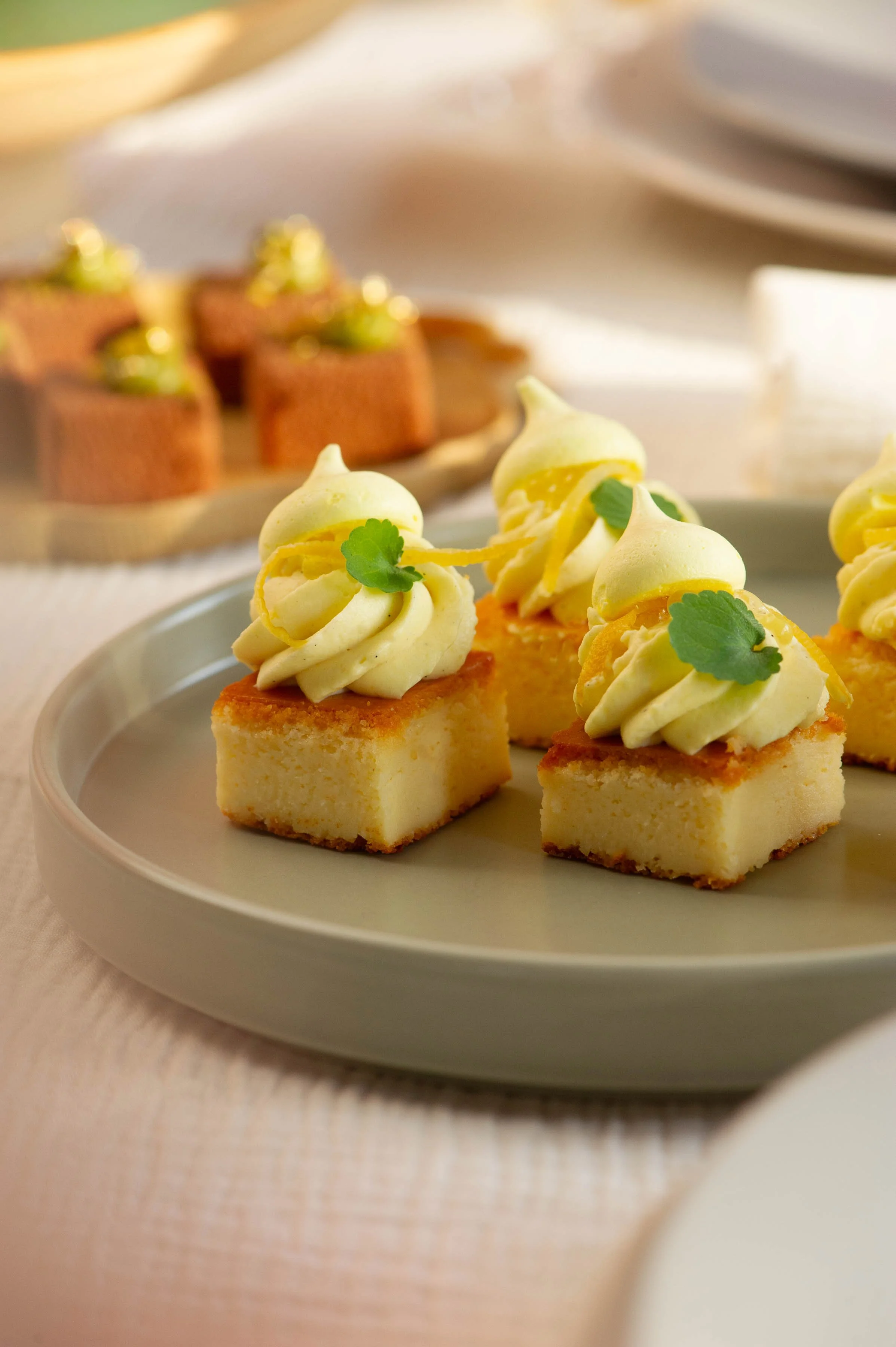 Dessert plate with small square cheesecake topped with whipped cream, lemon zest, and a mint leaf. Blurred background with additional cheesecake slices and a white cup.