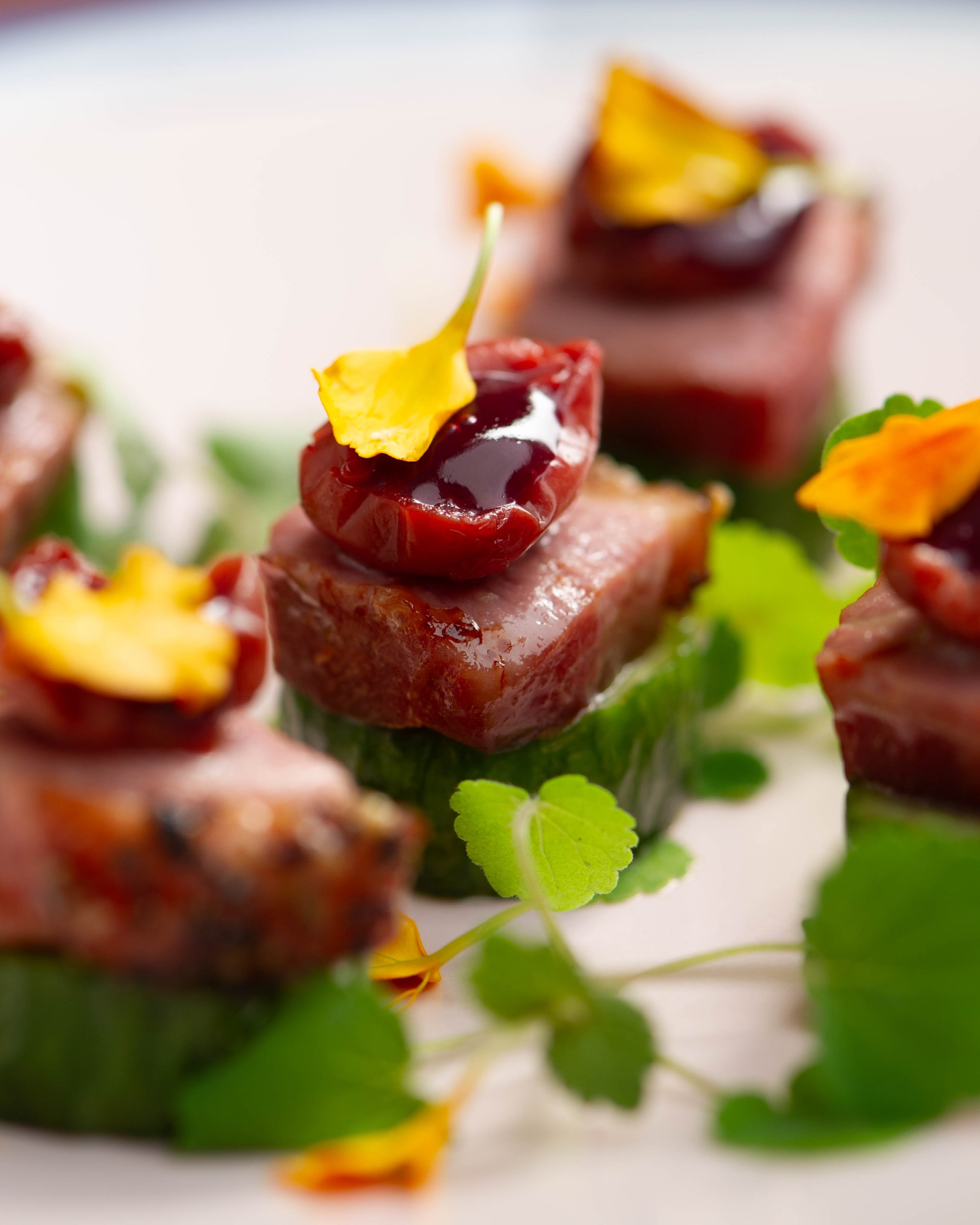 Close-up of gourmet appetizers with slices of red meat topped with dark cherry glaze and garnished with edible flower petals and green leafy herbs.