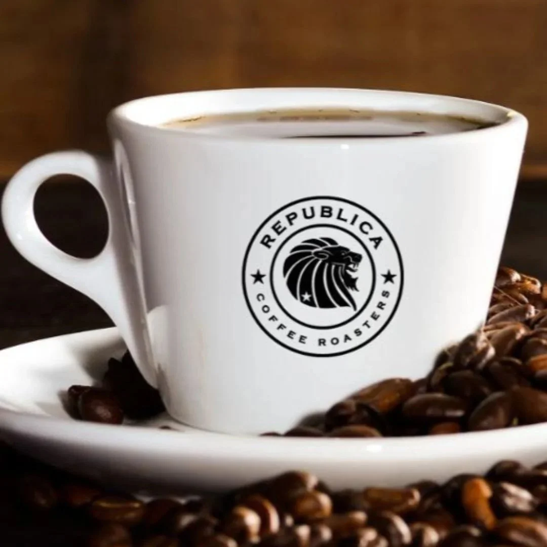 White coffee cup with 'REPUBLICA Coffee Roasters' logo, filled with coffee, placed on a saucer surrounded by coffee beans.
