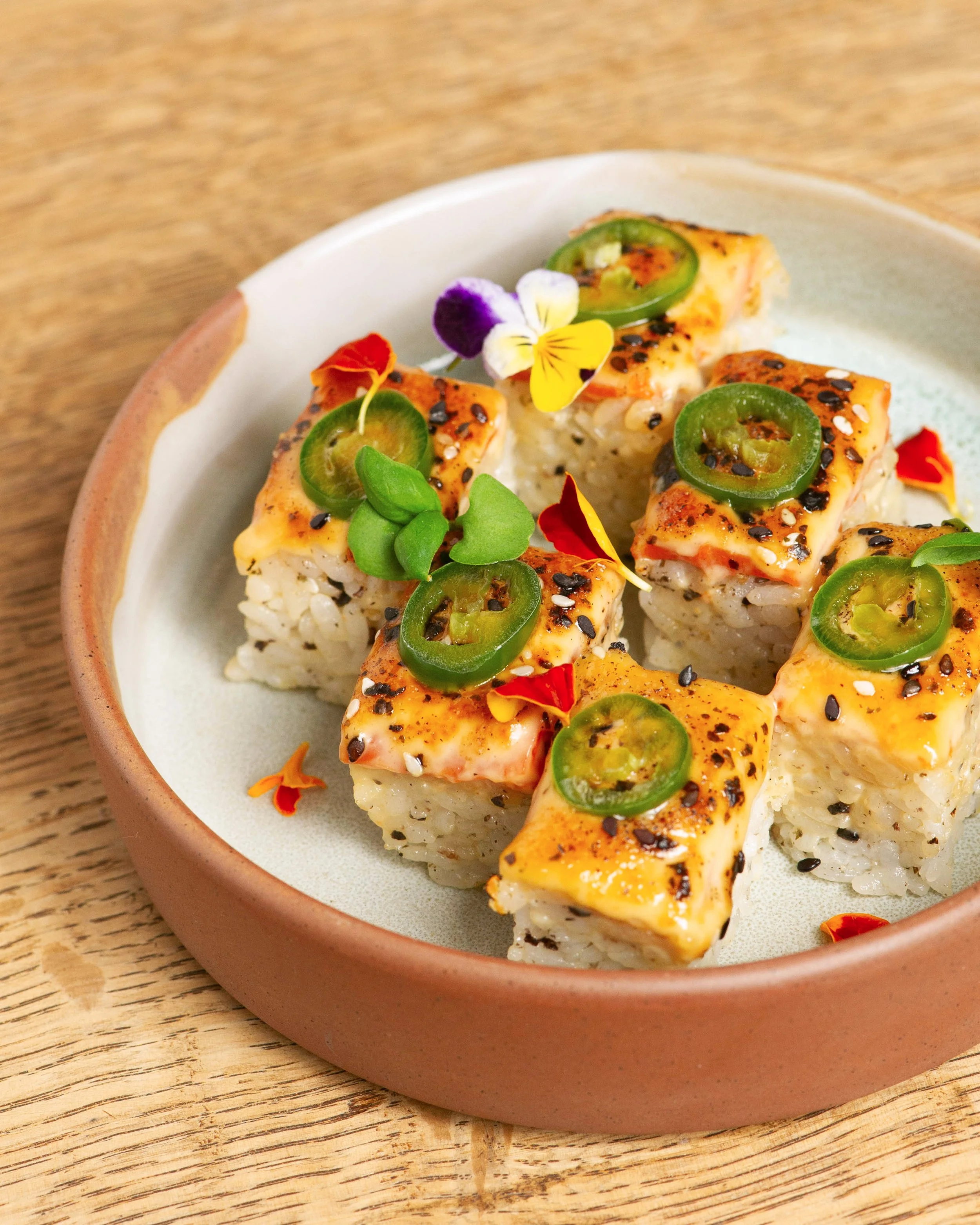 Close-up of sushi rolls topped with jalapeño slices, sesame seeds, and edible flowers on a ceramic plate, placed on a wooden surface.