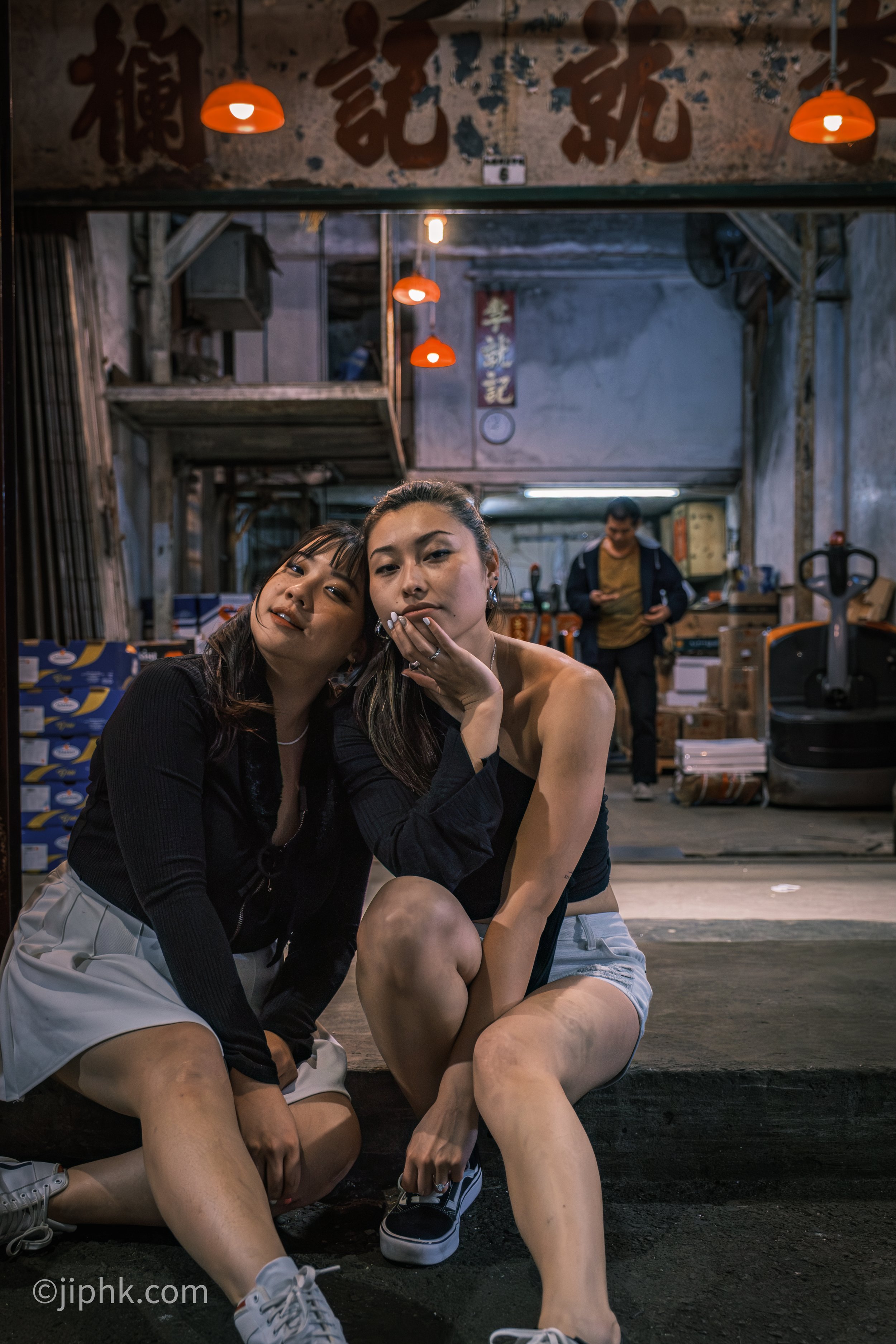 Two women sitting on a step in front of a market, posing for a photo at night with a man in the background inside the market.