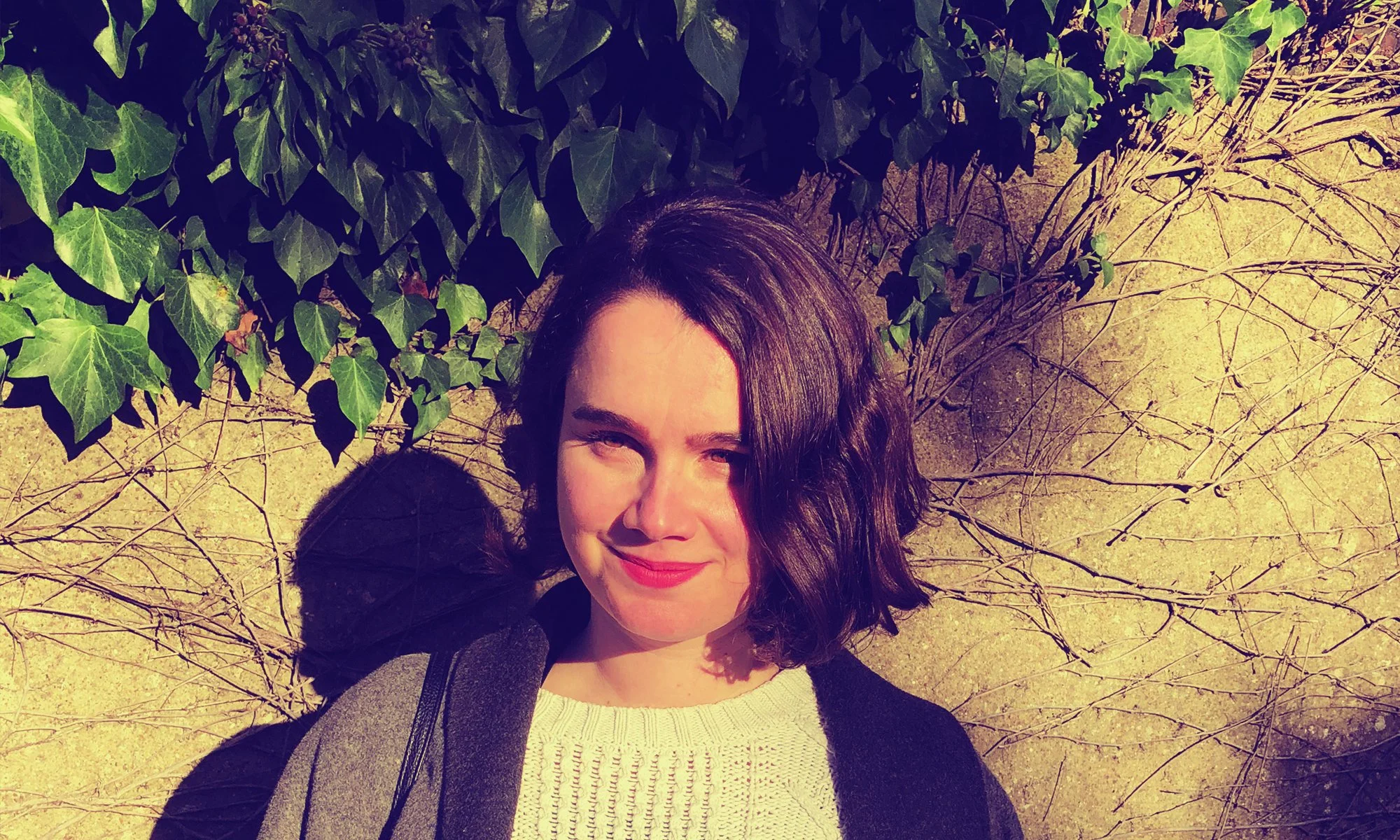 A photo of Annie Warburton looking off camera, wearing a white jumper and grey coat and standing in front of a wall with ivy growing on it.