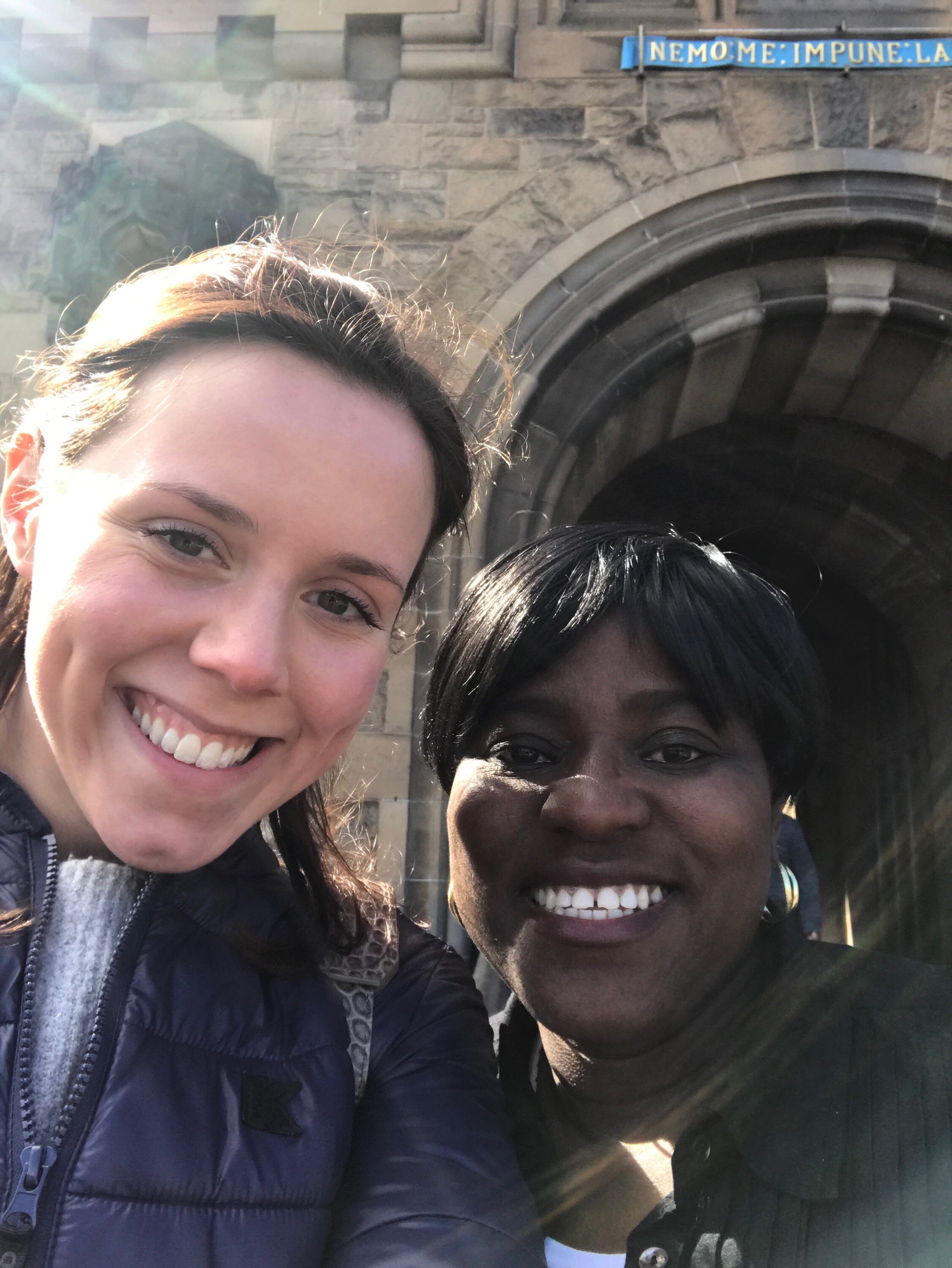 Two women smiling in front of an old stone archway, with sunlight creating lens flare.
