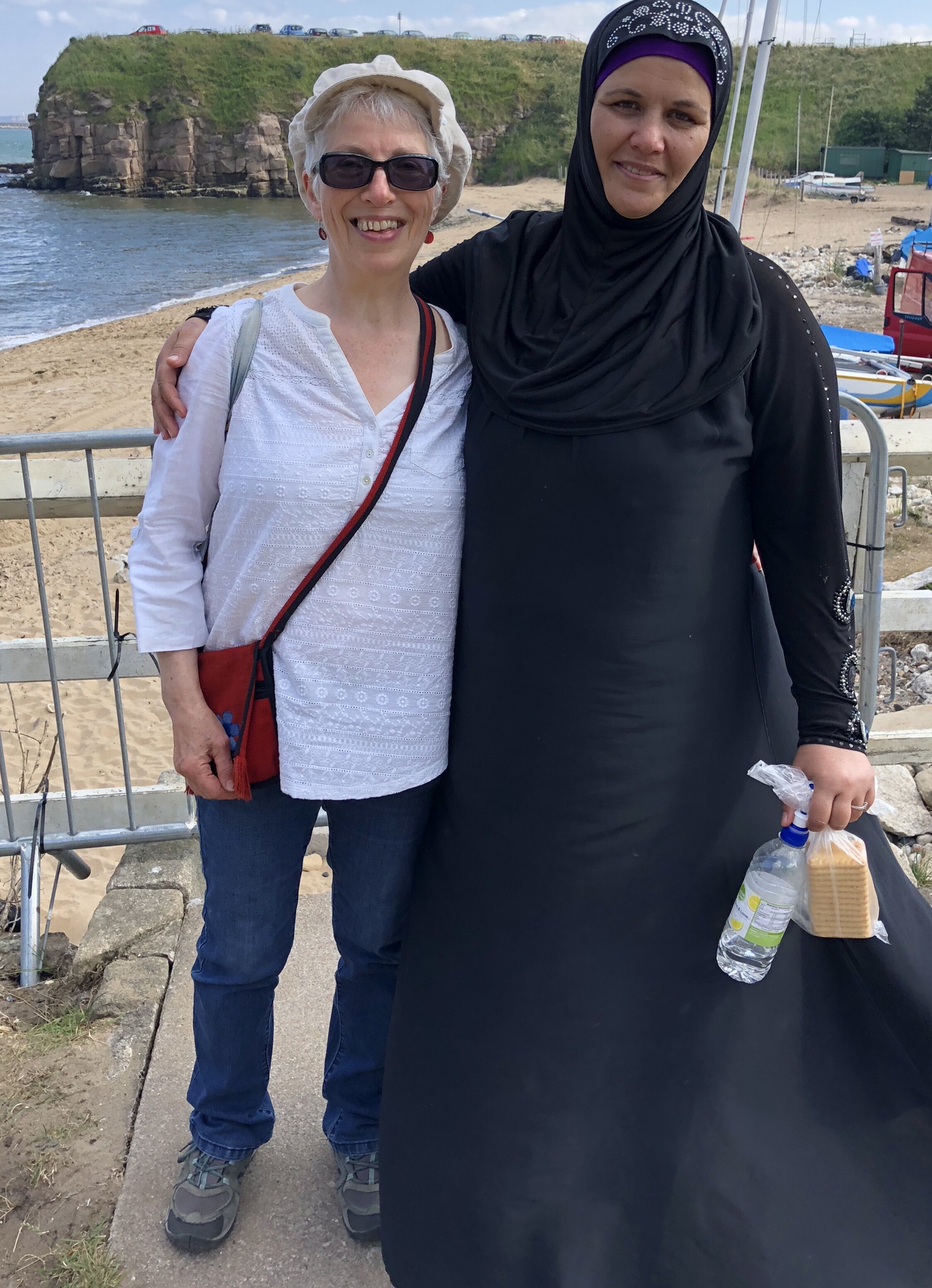 Two women standing on a beach path; one in a white blouse, sunglasses, and a white hat, the other in a black abaya and headscarf, holding a water bottle and biscuits. Background shows a rocky coastline and sandy beach.