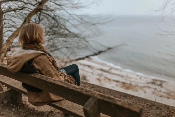 Woman sitting on bench by shoreline on gray day