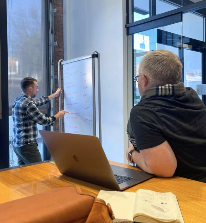 Two men in a modern office discussing written notes on a whiteboard. One man is sitting at a wooden table with a laptop and an open notebook. The other man, in a plaid shirt, is standing near the whiteboard, pointing at it.