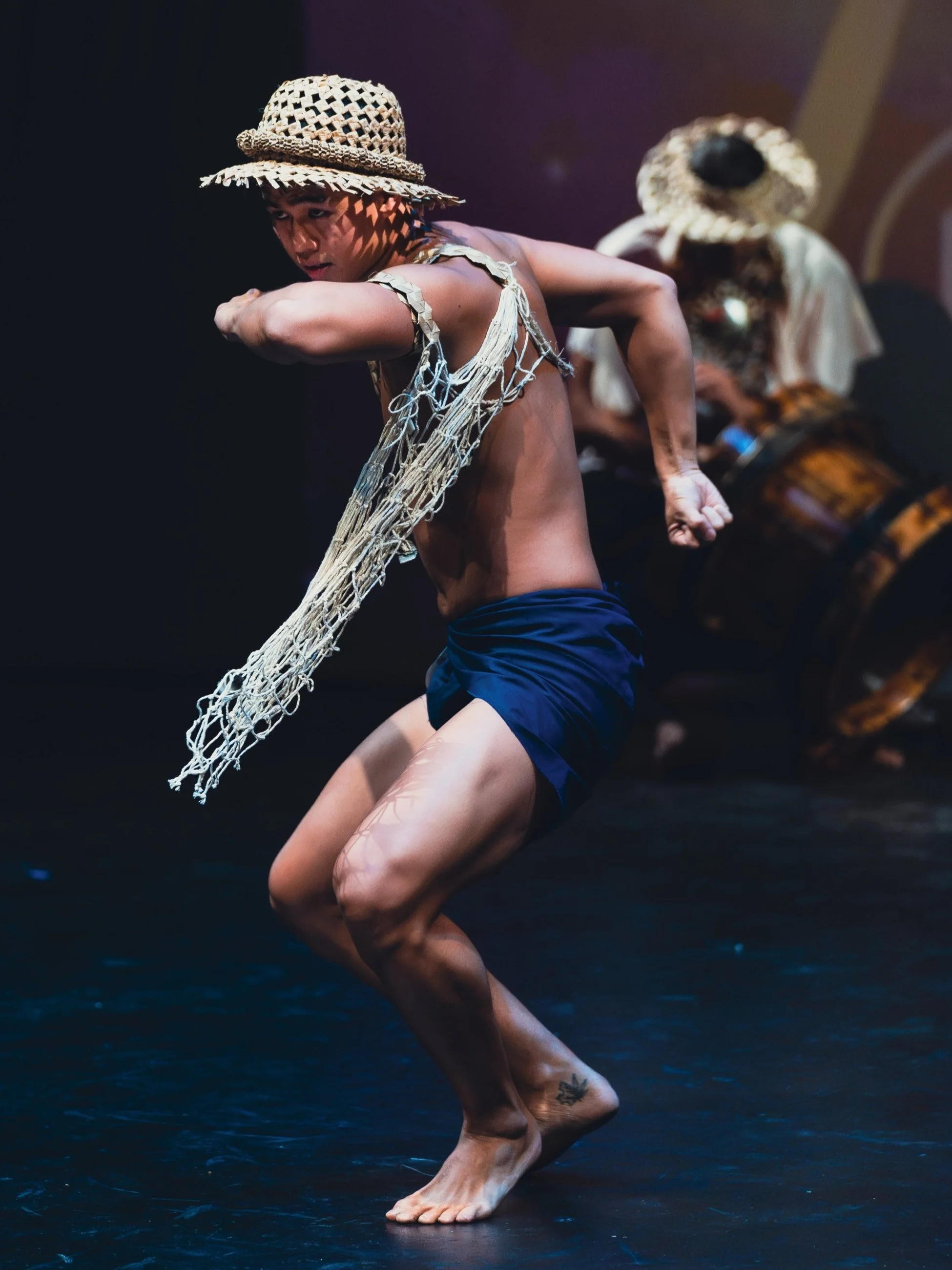 A dancer wearing a straw hat and a fringed costume performs energetically on stage, with a musician playing percussion in the background.