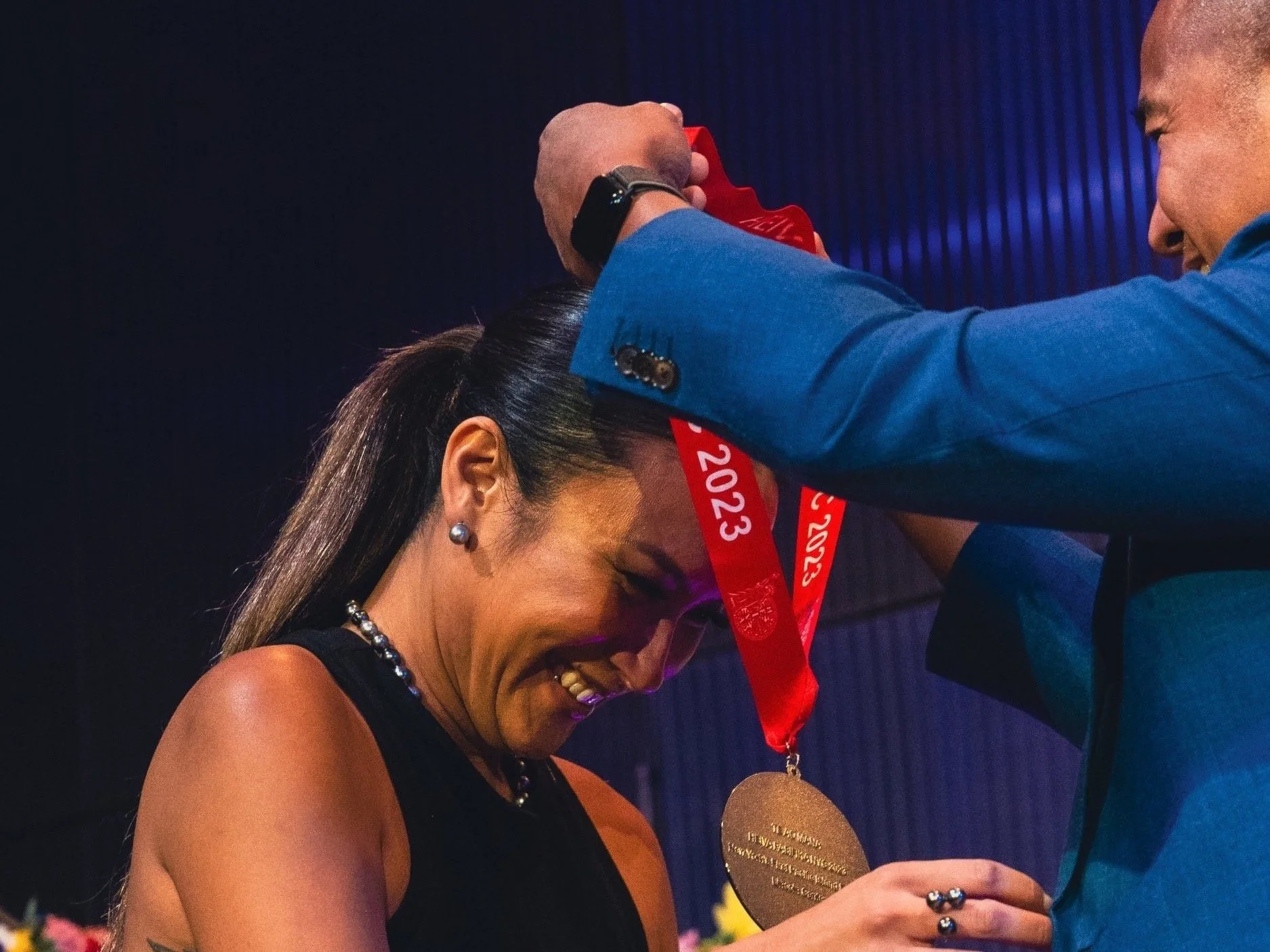 Person receiving a medal with the year 2023 engraved, placed around their neck by another person in a blue suit.