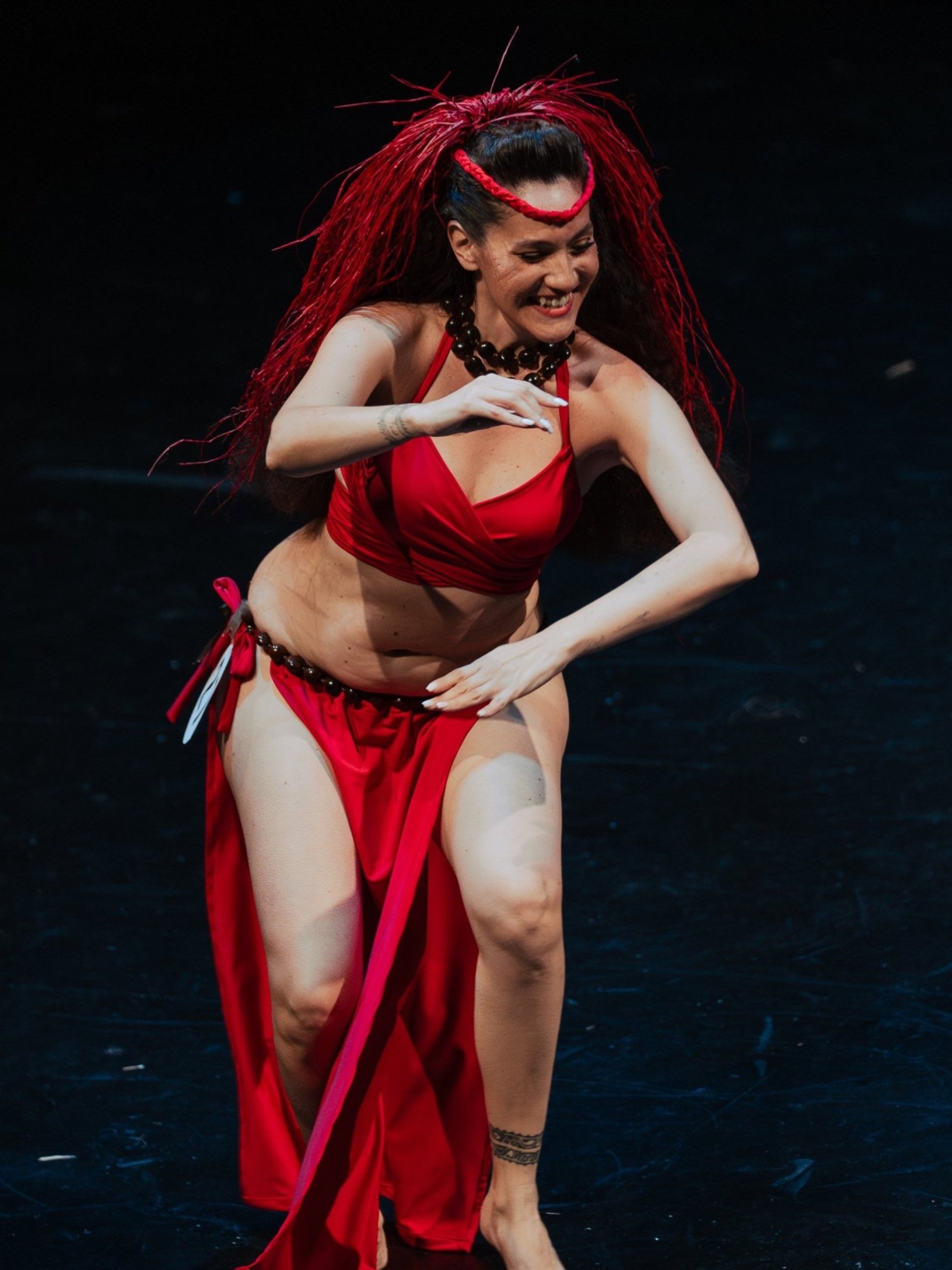Dancer performing in red costume on stage, wearing red headpiece and jewelry.