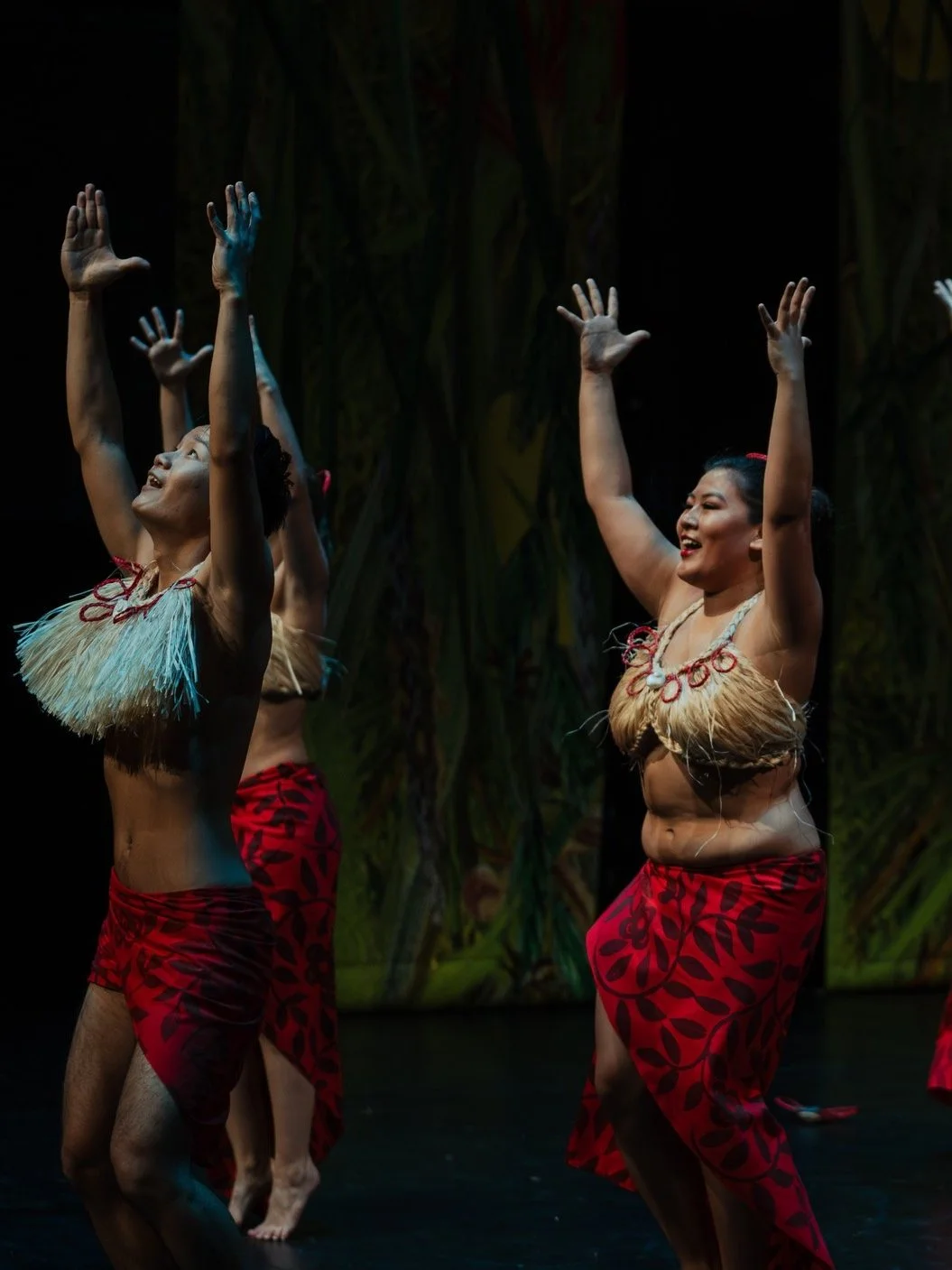 Traditional Polynesian dancers performing on stage in red costumes with straw tops, expressive hand movements and vibrant background.