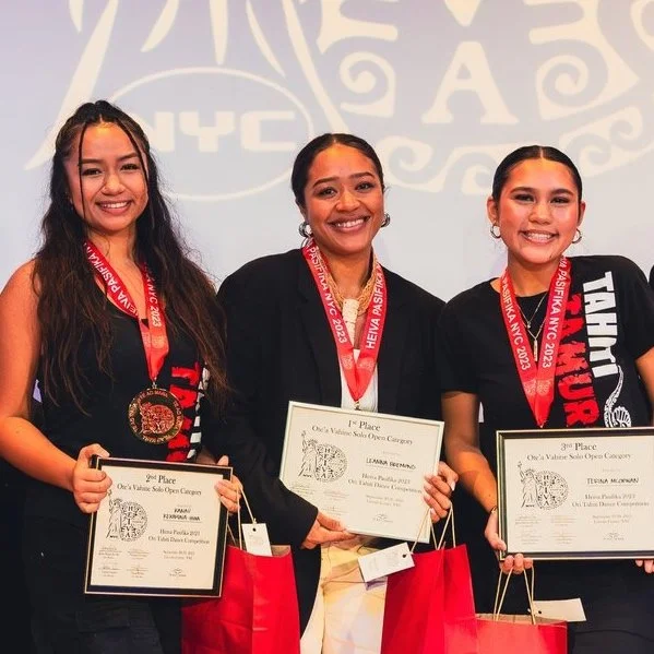 Three women holding certificates and wearing medals, smiling on stage. They have red ribboned medals around their necks and carry red shopping bags. Event branding is visible in the background.