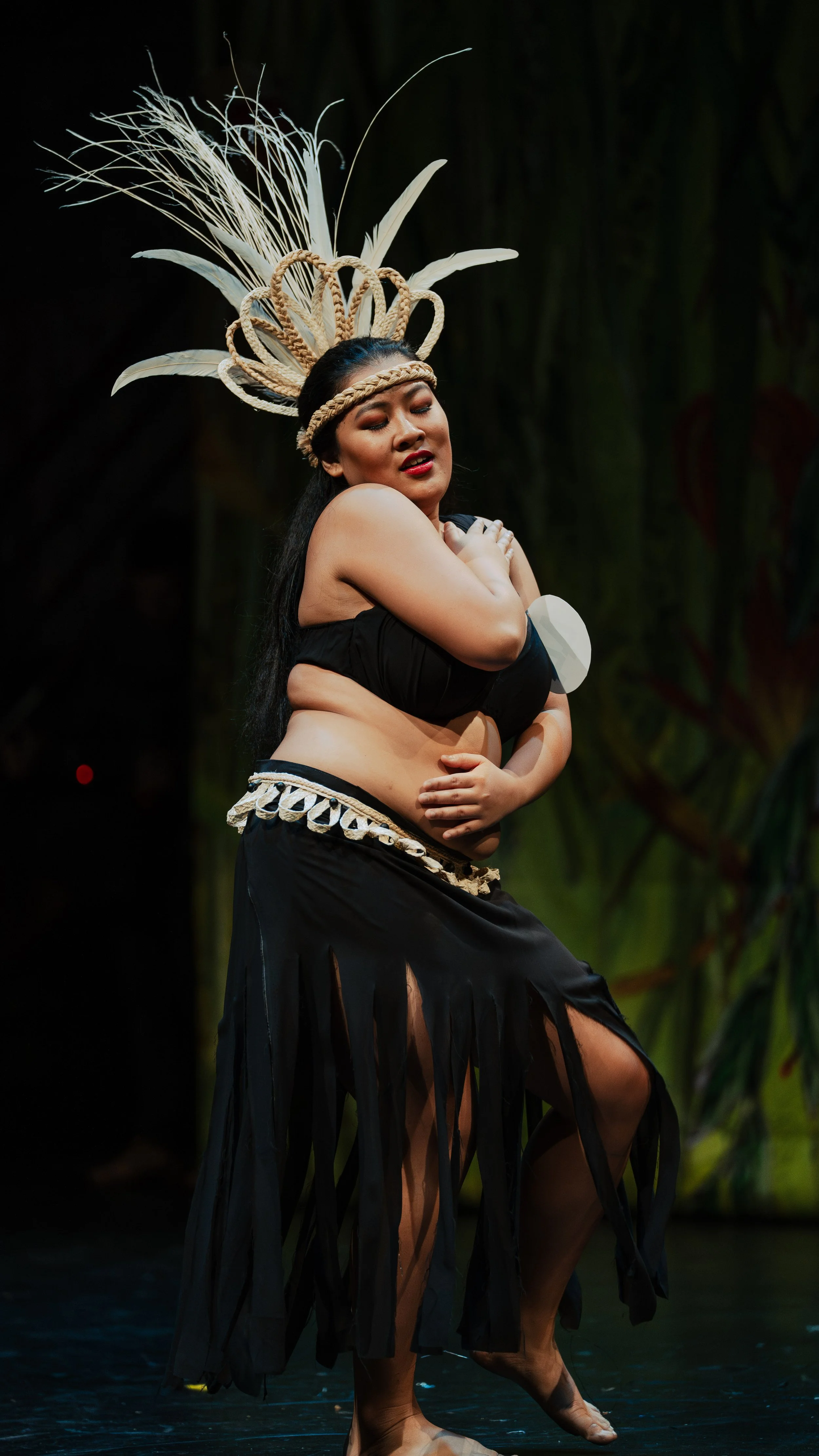 Performer dancing in traditional Polynesian attire with a headdress and black skirt on stage.