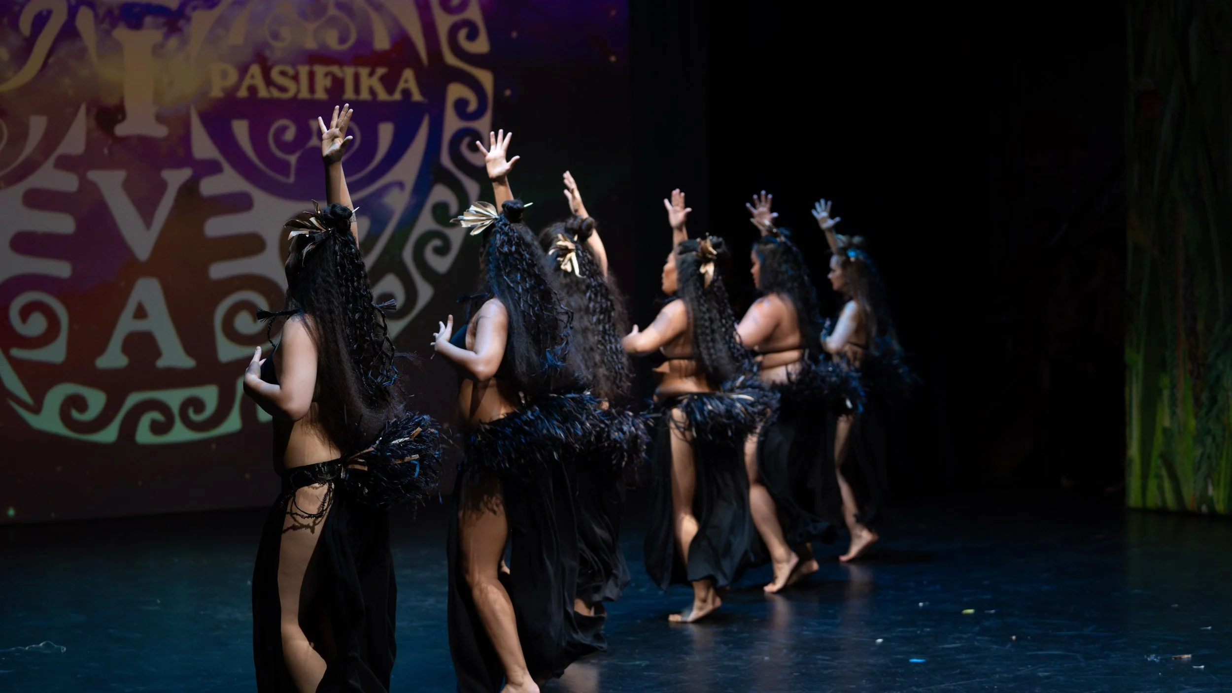 Group of women performing a traditional Polynesian dance on stage, wearing black skirts and feathered accessories.
